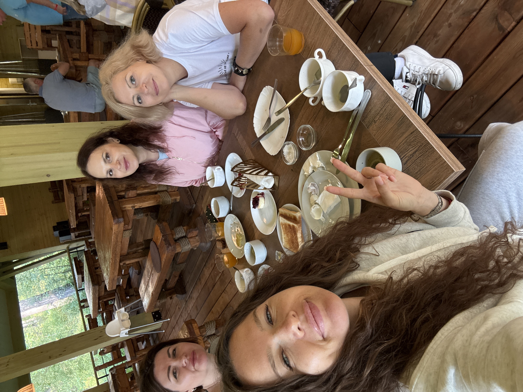 A group of four women are enjoying a meal together at an outdoor wooden restaurant in the city of Barangol, Russia. The restaurant appears to be in a forest setting, with trees visible in the background. The women are seated at a wooden table laden with various breakfast items, including toast, eggs, and fruit. They are all dressed casually and appear to be in good spirits, engaging in conversation and enjoying their meal. The overall atmosphere is relaxed and convivial.