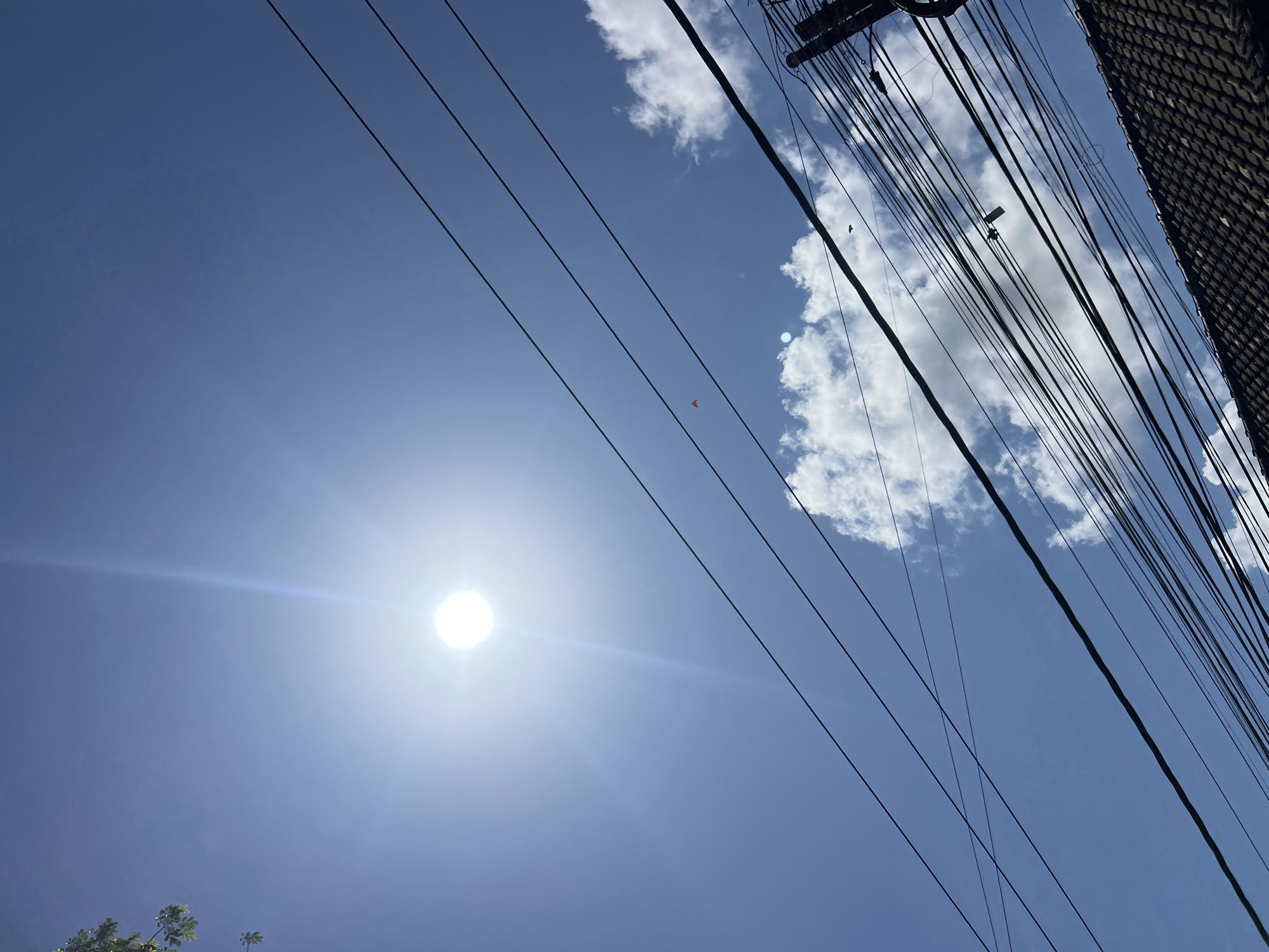 The image depicts a clear blue sky over Denpasar, Indonesia, with the sun prominently visible and creating a lens flare effect. Several electrical wires crisscross the frame, casting shadows and adding a sense of urban clutter. A small red kite is visible in the sky. In the foreground, the tiled roof of a building is partially visible, further grounding the scene in an urban environment. The presence of overhead wires and a tiled roof strongly suggests a residential or commercial area within the city.
