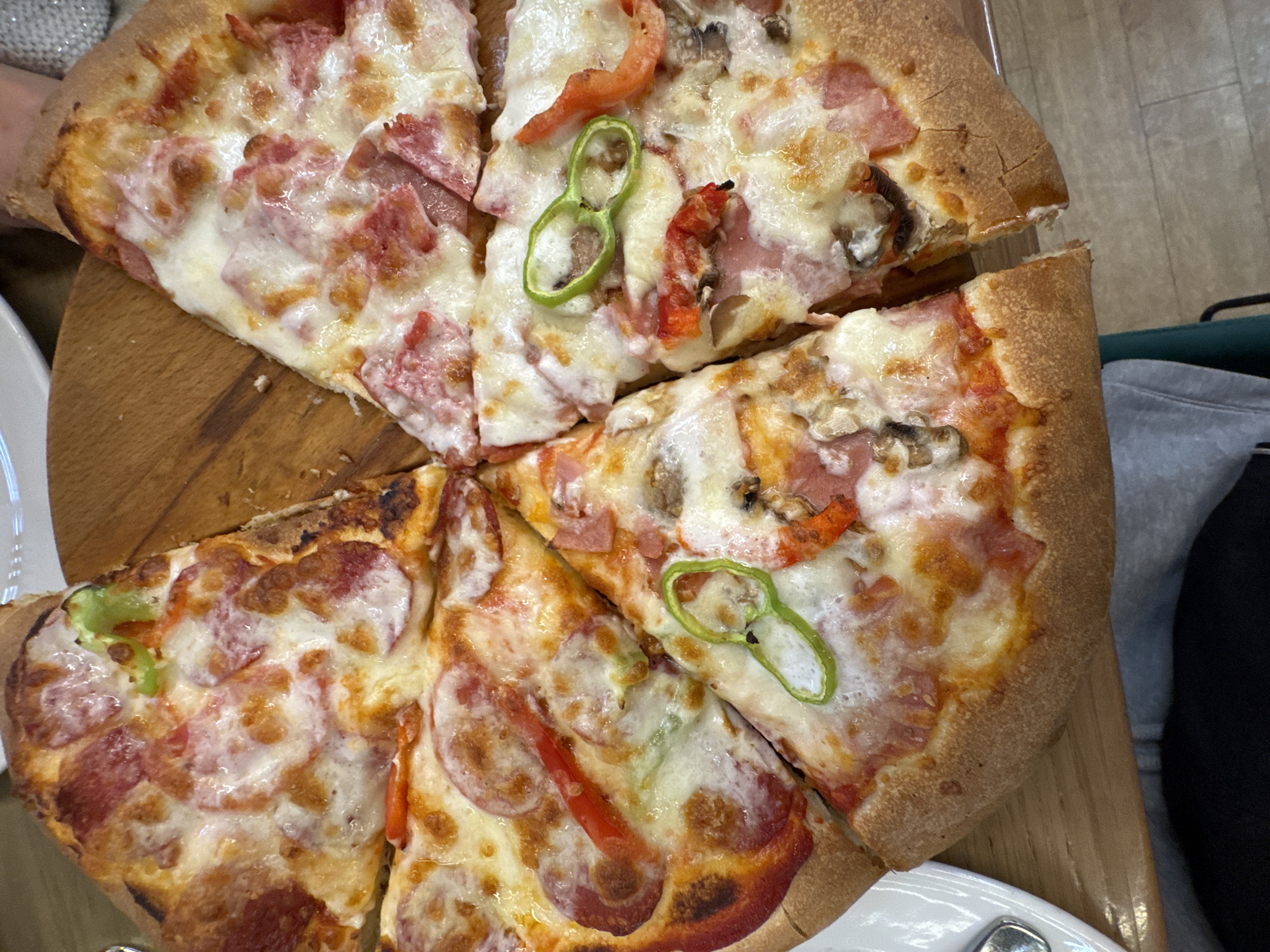 The image shows a partially eaten pizza served on a wooden platter. The pizza is cut into slices and appears to be topped with cheese, pepperoni, ham, tomatoes, mushrooms, and green pepper rings. The pizza has a thick crust. The platter sits on a wooden table. Background details include parts of a white plate, a light gray sweatshirt, and possibly a green bag. The location appears to be indoors, possibly a restaurant. Information on Yerevan, Armenia is not provided in the image.