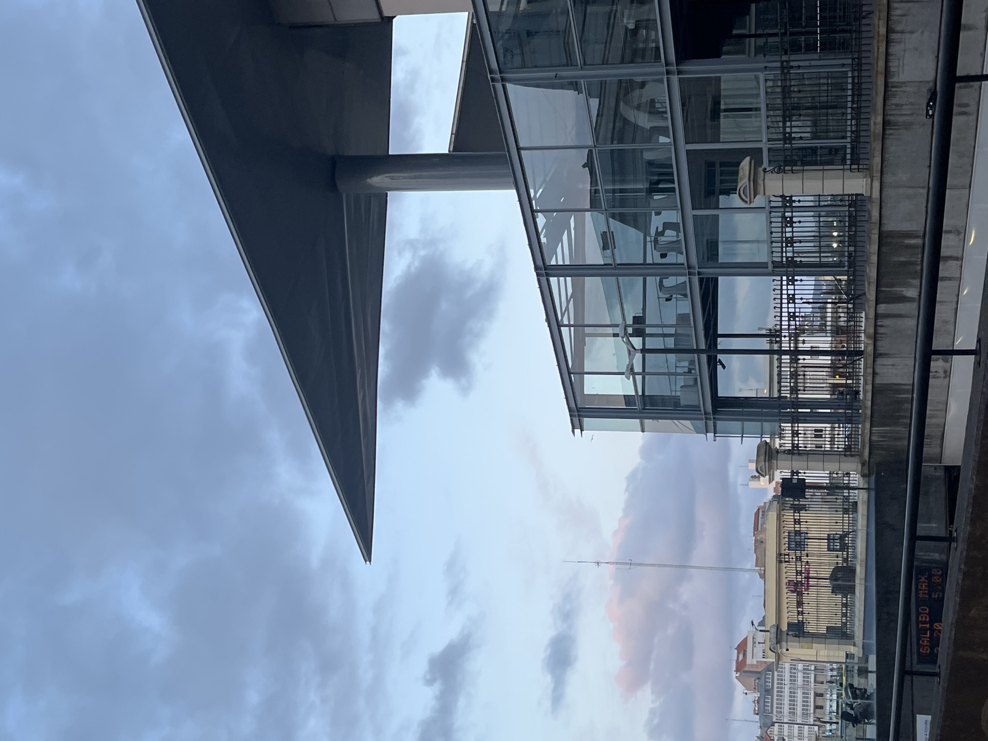 The image shows a partial view of a modern glass and steel building in La Coruña, Spain, taken from a low angle. A large, triangular grey overhang is visible on the left, supported by a cylindrical column. Adjacent to it is a multi-story glass structure with a visible interior, reflecting the exterior environment. Iron railings separate the building from the foreground. The background shows a cityscape with older buildings, their facades visible through the reflections and glass of the modern structure. The sky is overcast with grey and pink-tinged clouds. The image has been rotated 90 degrees counterclockwise.