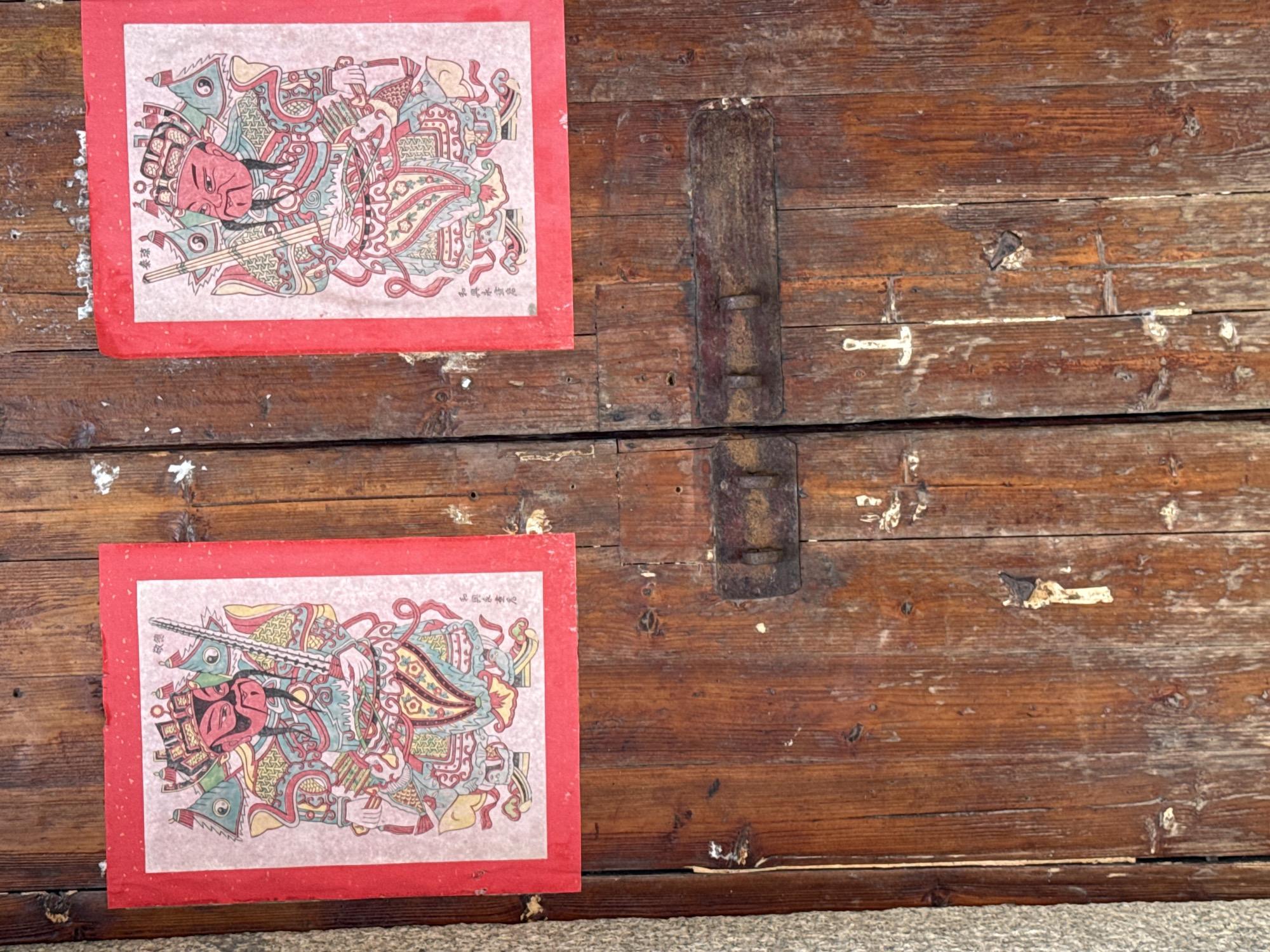 The image shows a close-up of a weathered wooden double door with two red-bordered New Year's prints adhered to the upper panels. The prints, depicting traditional Chinese figures in armor, appear to be the same image on both doors. The figures are facing forward and holding weapons, with red faces and elaborate headdresses. Chinese characters are visible on the prints. Below the prints, a rustic, tarnished metal latch assembly is attached horizontally across the seam of the doors. The wood of the door is aged, with visible grain, knots, and some peeling paint or discoloration. The overall impression is of an old, traditional entryway, possibly in a residential or older commercial building. The location is specified as Longgang District, China.