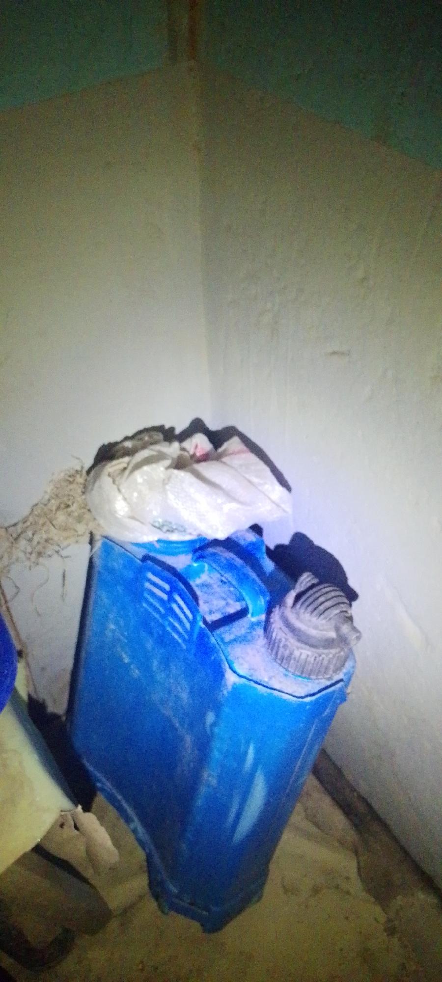 The image depicts a blue plastic jerrycan in a room corner. The jerrycan appears to be dirty and dusty, with a white bag or packaging placed on top of it. The walls of the room are painted a light color, and there is some debris on the floor. The lighting in the image is dim, with a single light source creating shadows. The scene suggests a storage area or a neglected space. It is not possible to determine the specific location within Jalingo, Nigeria, from the image alone.