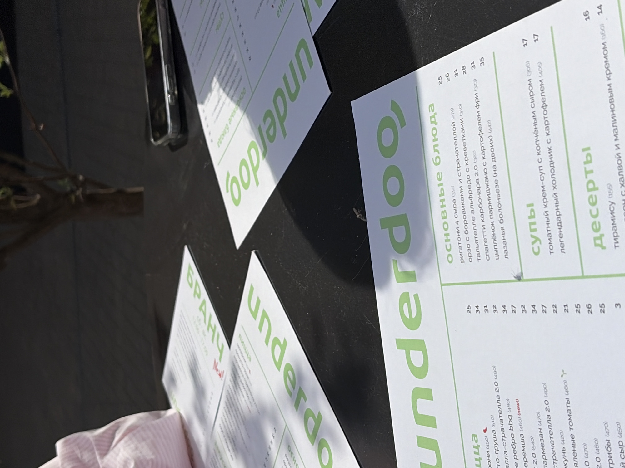 The image shows a table in a presumably outdoor setting, possibly in Minsk, Belarus, due to the presence of text in Russian. Various menus with the restaurant name "underdog" are visible, with "БРАНЧ" (Brunch) on one. The menus detail food items with prices next to them, including pizza, main courses, soups, and desserts.