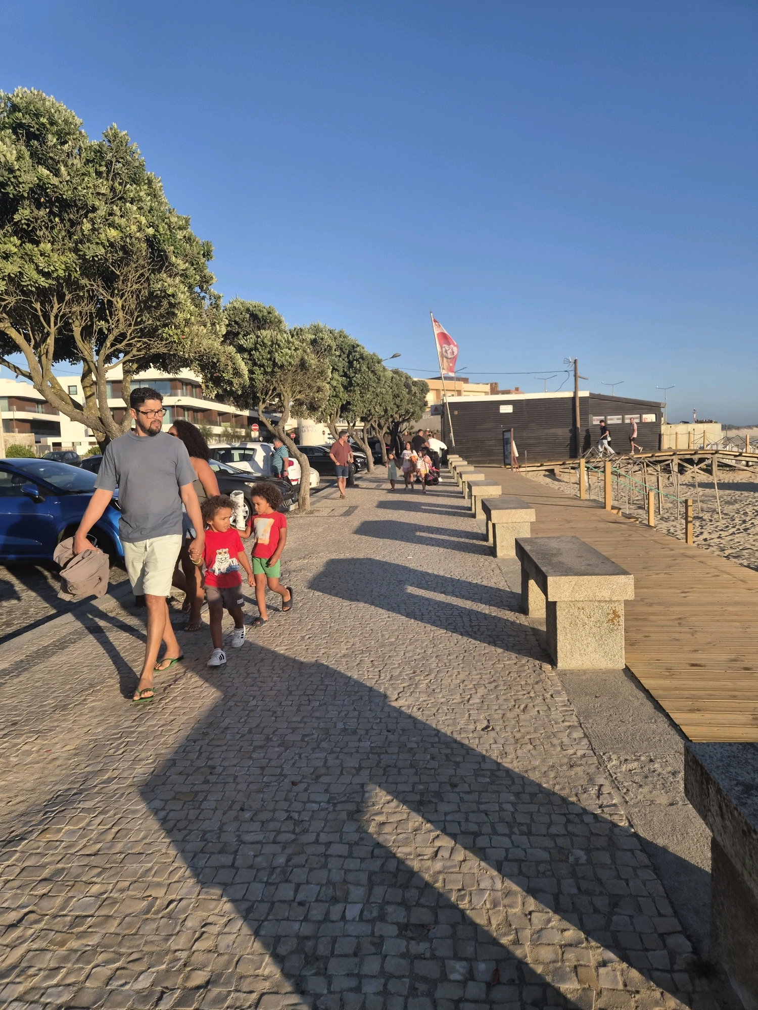 The image shows a sunny day with a clear blue sky in Esposende, Portugal. A paved walkway with stone benches lines the right side of the image, bordering a sandy beach. A wooden boardwalk runs parallel to the beach. Several people are visible walking along the paved path. In the foreground, a man in a blue t-shirt and light shorts walks with two young children, a boy in a red t-shirt and shorts, and another boy in a red shirt and green shorts. They are walking away from the camera. In the background, a dark wooden building with a red and white flag is visible. Trees with green foliage line the path between the walkway and the buildings. Cars are parked along the left side of the path.