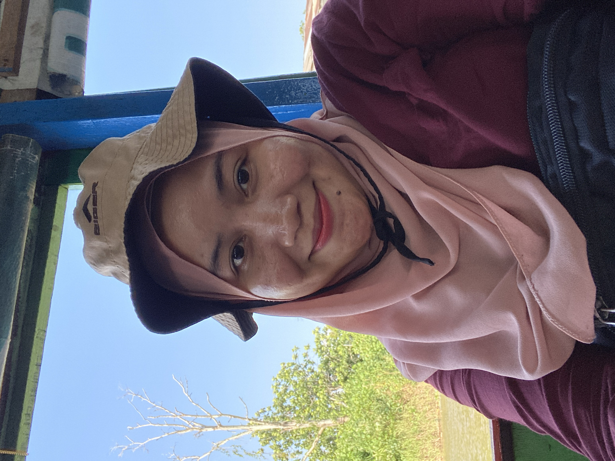 A woman wearing a pink hijab and a beige bucket hat with the brand "Eiger" is smiling at the camera. She is wearing a maroon-colored shirt. She appears to be on a boat, with a body of water and lush green vegetation visible in the background. The sky is clear blue. The overall scene suggests a casual outing in a natural environment, possibly a river tour in or near Banjarmasin, Indonesia, given the presence of water and dense foliage.