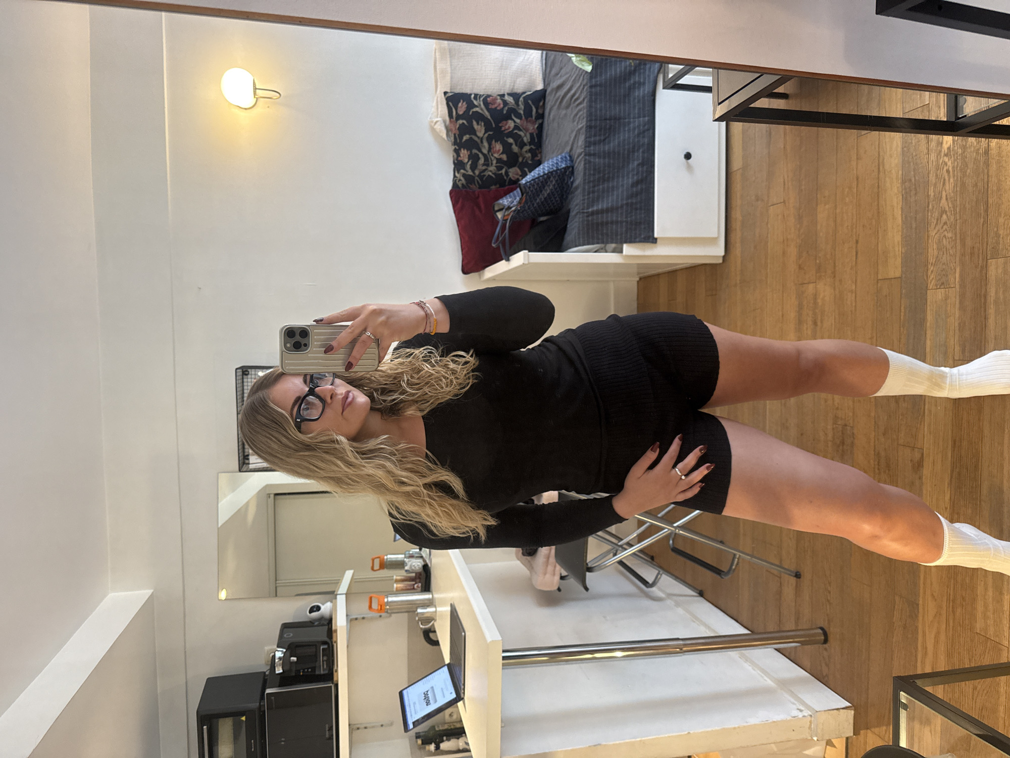 A young woman wearing a black long-sleeved dress and white socks is taking a mirror selfie in what appears to be an apartment. She is holding a smartphone, and her reflection shows a room with a bed, pillows, and a table with a glass top. In the background, a kitchenette area is visible with appliances and various items. The apartment has white walls and wooden floors. The scene suggests a modern, lived-in space that is likely located in a residential area in an urban setting, possibly in Paris, France.