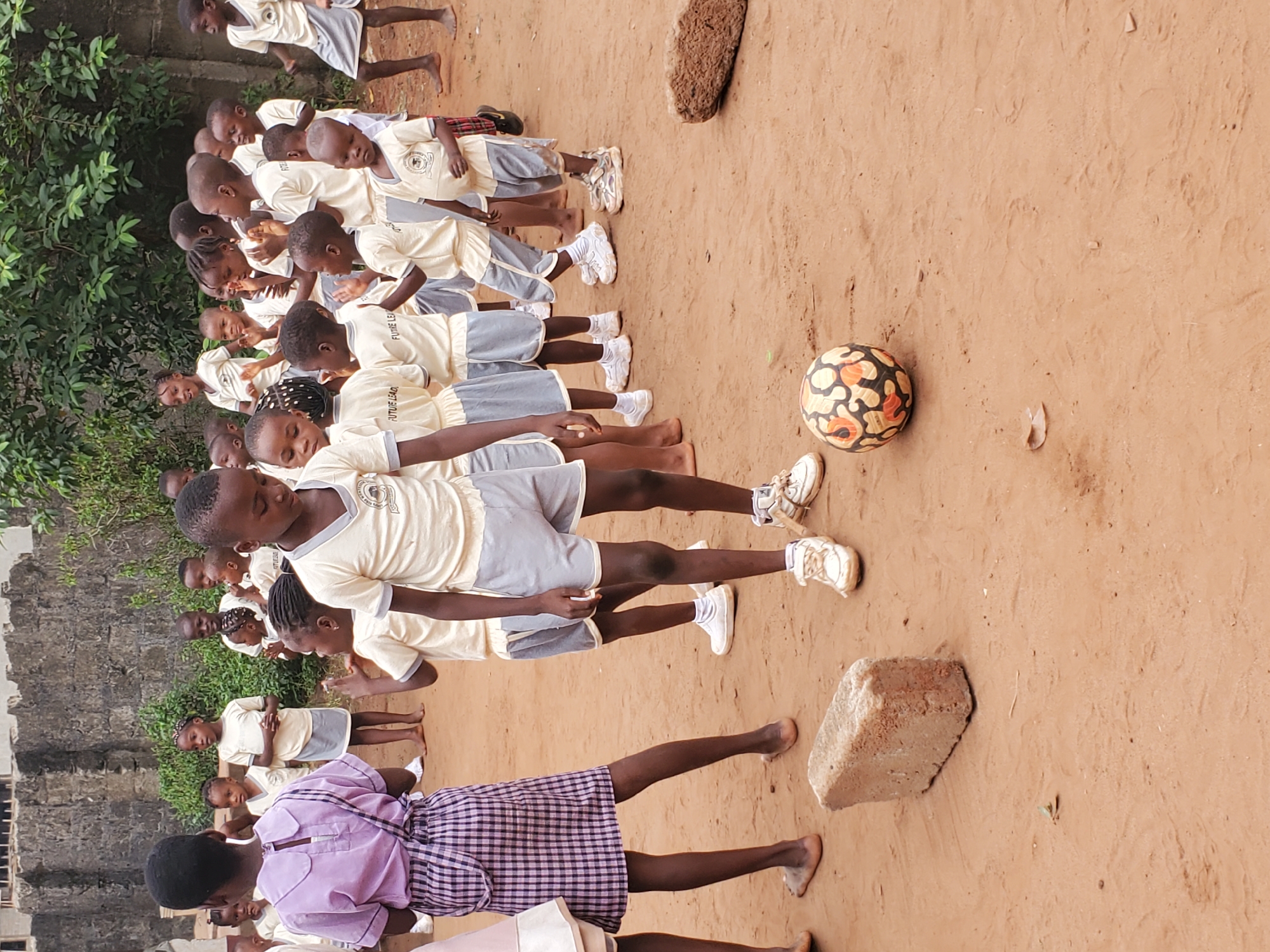A group of children wearing matching yellow shirts and grey shorts stand in a line in an outdoor area in Ukwu-Abwa, Nigeria. Most of the children are wearing white sneakers. The ground is a reddish-brown color. A soccer ball sits to the side of the line. There are two rocks on the ground as well. There is a brick building and green foliage in the background. One child at the front of the line is wearing a pink shirt and a blue and white checkered skirt, and no shoes.