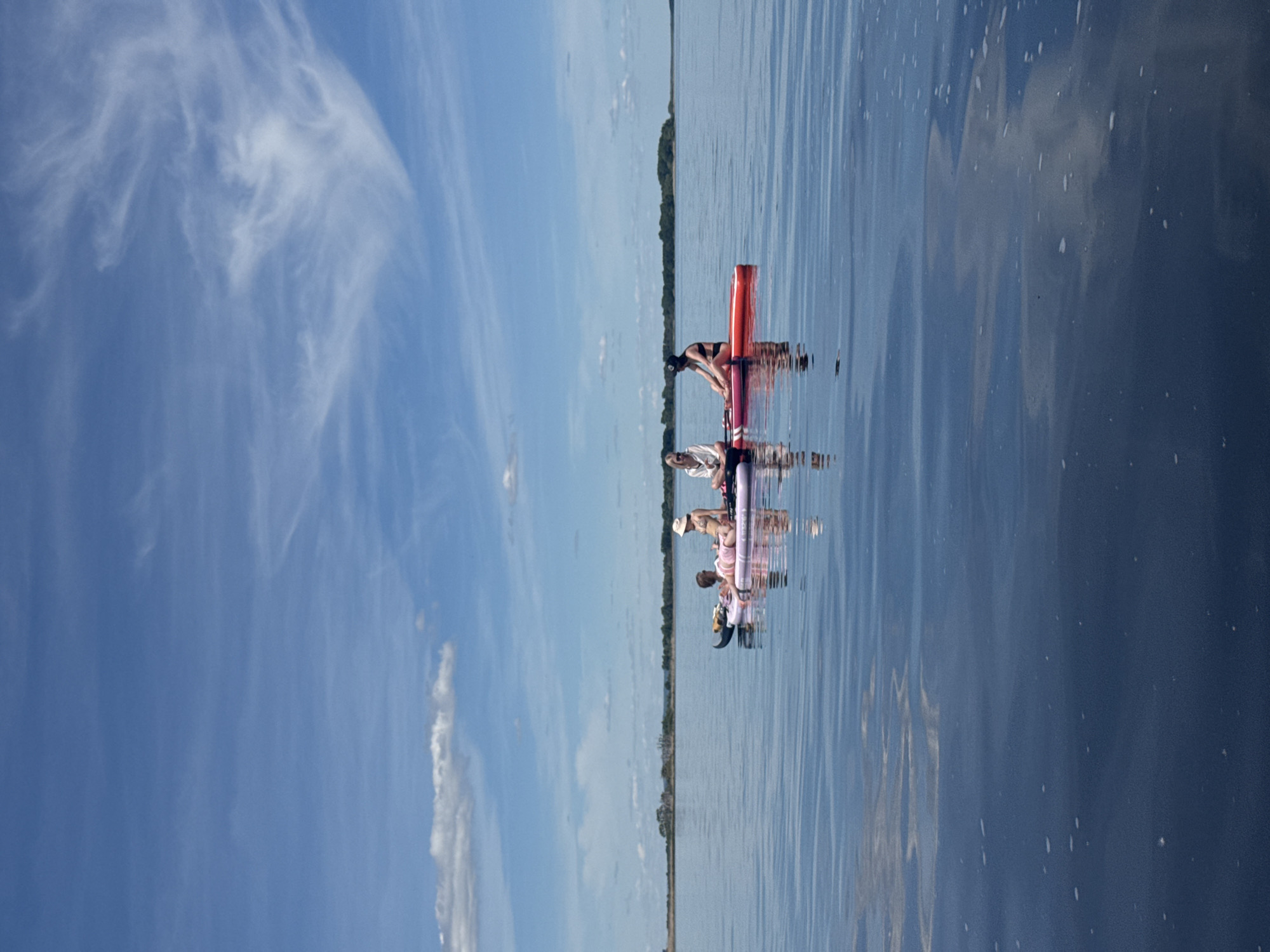 The image displays a serene outdoor scene, correctly oriented in a landscape format, featuring a group of people enjoying a calm body of water under a clear blue sky. The setting is likely a wide river or lake, consistent with the provided context of Усть-Курдюм, Russia.
The sky occupies a significant portion of the upper frame, a vibrant blue adorned with delicate, wispy white clouds (cirrus clouds), suggesting fair and pleasant weather. Below the sky, the horizon is marked by a distant, low-lying treeline or landmass, indicating the expansive nature of the water body.
The water itself is tranquil and highly reflective, mirroring the blue of the sky and the white clouds, creating subtle patterns and slight ripples on its surface. Several individuals are visible on the water, appearing to be engaged in recreational activities such as paddleboarding. In the foreground-middle ground, a person in a dark swimsuit or top is kneeling on a bright red inflatable paddleboard. Behind them, another person is situated on a purple or pink inflatable board. Further in the distance, additional figures can be seen on similar watercraft, forming a small group enjoying the aquatic environment.
The time of day appears to be bright daylight, possibly mid-morning or early afternoon, given the clear visibility and the quality of light. The overall atmosphere conveyed is one of peaceful recreation and enjoyment of nature. There is no visible text identifiable on any objects or in the environment.