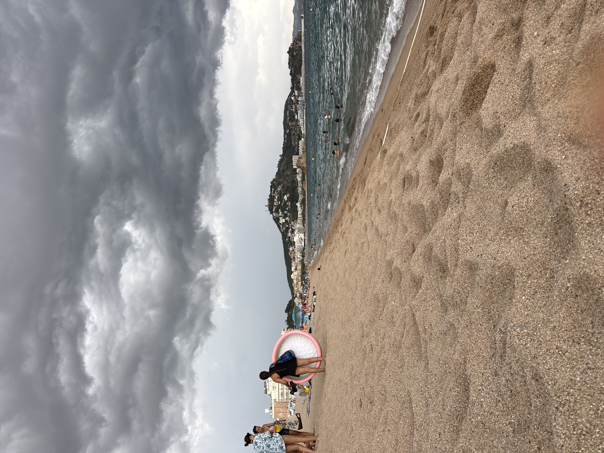 The image captures a view of the beach in Blanes, Spain, under a heavily overcast sky with dark, dramatic clouds dominating the upper portion of the frame. In the foreground, a sandy beach stretches towards the sea.
On the left side of the beach, three individuals are visible. Two women stand closer to the camera, one wearing a white and blue patterned shirt and shorts, and the other in a black swimsuit. The woman in the swimsuit is holding a large, inflatable pink ring. A third person, partially obscured, is also present.
Further down the beach, the sand transitions to the sea. Several people are in the water, swimming or wading. The sea is a deep blue-green, with gentle waves lapping at the shore. In the distance, the town of Blanes is spread out along the coastline, with buildings climbing up a hillside. A prominent structure, possibly a castle or watchtower, is visible on the highest point of the hill. The overall atmosphere suggests a cloudy day, possibly with a threat of rain, but people are still enjoying the beach and the water.