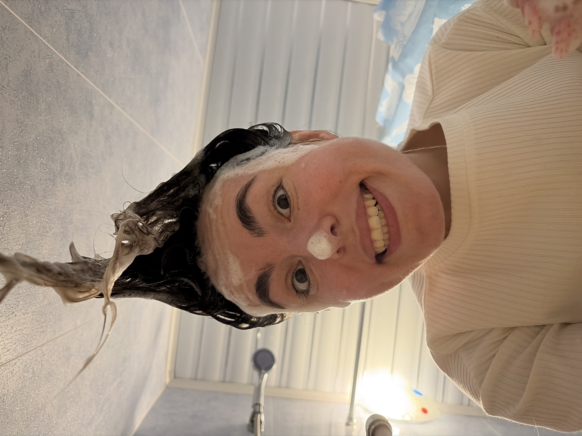 A woman with dark, wet hair, lathered with soap on her face and hair, is looking down at the camera, smiling. She is wearing a white, ribbed shirt. She is in a shower with white tiled walls and a blue accent wall. The shower head and fixtures are visible below her. This image does not provide information to determine the location of Izhevsk, Russia.