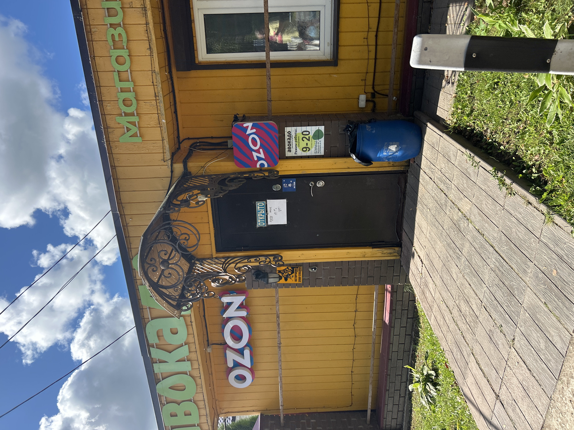 The image shows the exterior of a brightly painted yellow building, likely a small shop or kiosk, located in Kalitsino, Russia. A wooden walkway leads to a dark-colored door. Above the door, a decorative metal awning is visible. A prominent "OZON" logo, with white text on a pink and blue striped background, is mounted on the wall to the right of the door. Another sign with "OZON" in white text is visible on the upper left portion of the building. To the right of the door, a sign displays "АВОКАДО" (Avocado) in green text, with operating hours of "9-20" (9 AM to 8 PM) below it. A blue barrel sits next to the door. The building's facade is adorned with green lettering that reads "АВОКАДО" on the left and "МАГАЗИН" (Shop) on the right, both in large, raised letters. A single window is visible on the right side of the building, through which a person wearing a yellow garment can be seen. The area is outdoors, with green grass and some foliage surrounding the building. The sky above is blue with scattered white clouds. A black and white striped post stands in the foreground to the right.