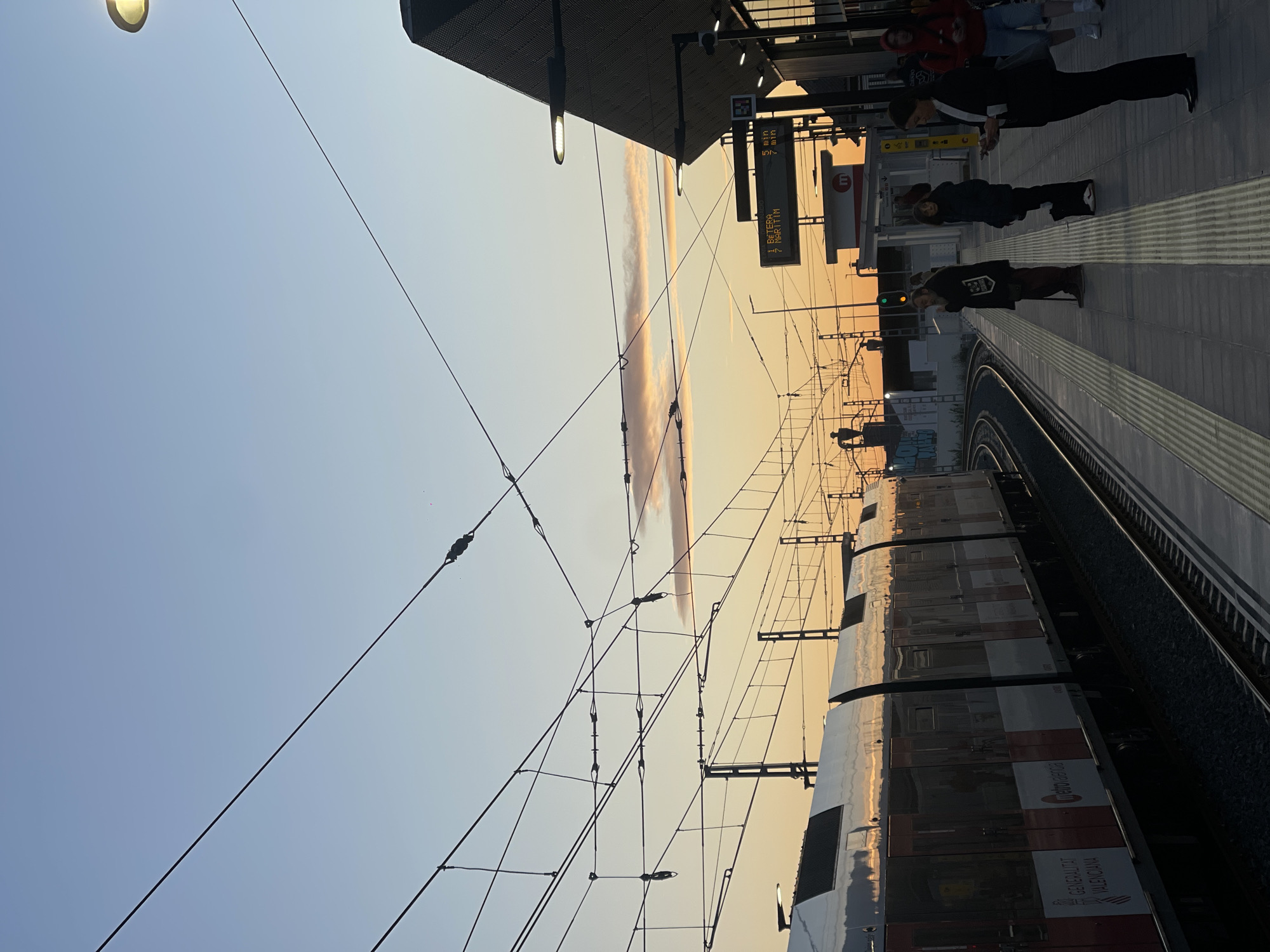 The image depicts a train station platform in Paiporta, Spain. A train is visible, bearing the markings "Metro Valencia" and "Generalitat Valenciana." Above, a sign indicates travel towards "I. Betera" and "7 Maritim." Passengers are standing on the platform. The sky is tinted with the colors of sunset. Overhead wires are present.