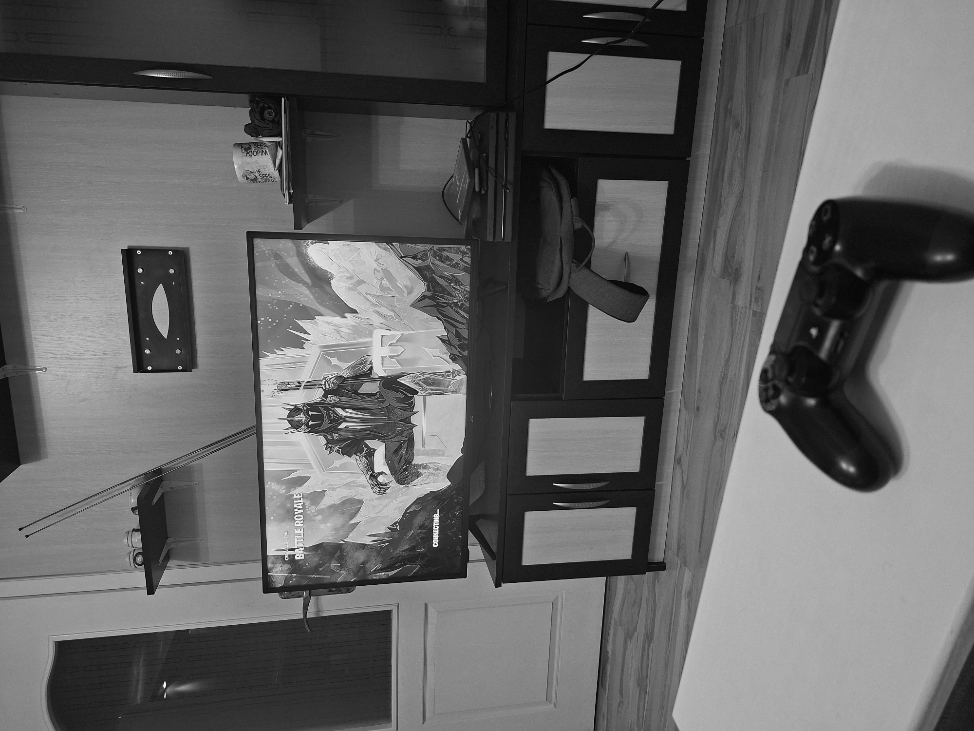 A grayscale, rotated image presents a room interior. A television screen displays the words "BATTLE ROYALE" and "CONNECTING..." with an image of a warrior figure. To the right of the screen is a black cabinet with three drawers, each featuring a wooden panel. Above the cabinet is a shelf with a PlayStation console. A backpack hangs from the side of the cabinet. A black PlayStation controller rests on a white surface to the right of the cabinet. An antenna and a wall mount bracket are visible on the wall to the left of the screen. A window is positioned below the screen. The image orientation suggests it has been rotated counterclockwise. The location in Plovdiv, Bulgaria is not discernible from the image.
