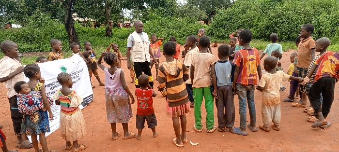 The image depicts an outdoor scene with approximately 25 to 30 children and one adult male in Ngalula, Democratic Republic of the Congo. The ground is reddish-brown dirt, and the background consists of dense green foliage and trees. An adult male, wearing a light-colored multi-pocket vest over a blue shirt and dark trousers, stands towards the left-center, facing the children. On the far left, several children, one with an infant secured in a patterned fabric carrier on their back, hold a white banner horizontally. The banner displays text, including phrases such as "POUR LES ENFANTS DE RESSORTISSANTS..." and "DRC," along with a green circular logo. The majority of the children, ranging in age from toddlers to early teenagers, are positioned in a loose semi-circular formation, predominantly on the right side, with many holding hands. They are dressed in various casual clothing; some are barefoot, while others wear sandals or flip-flops.