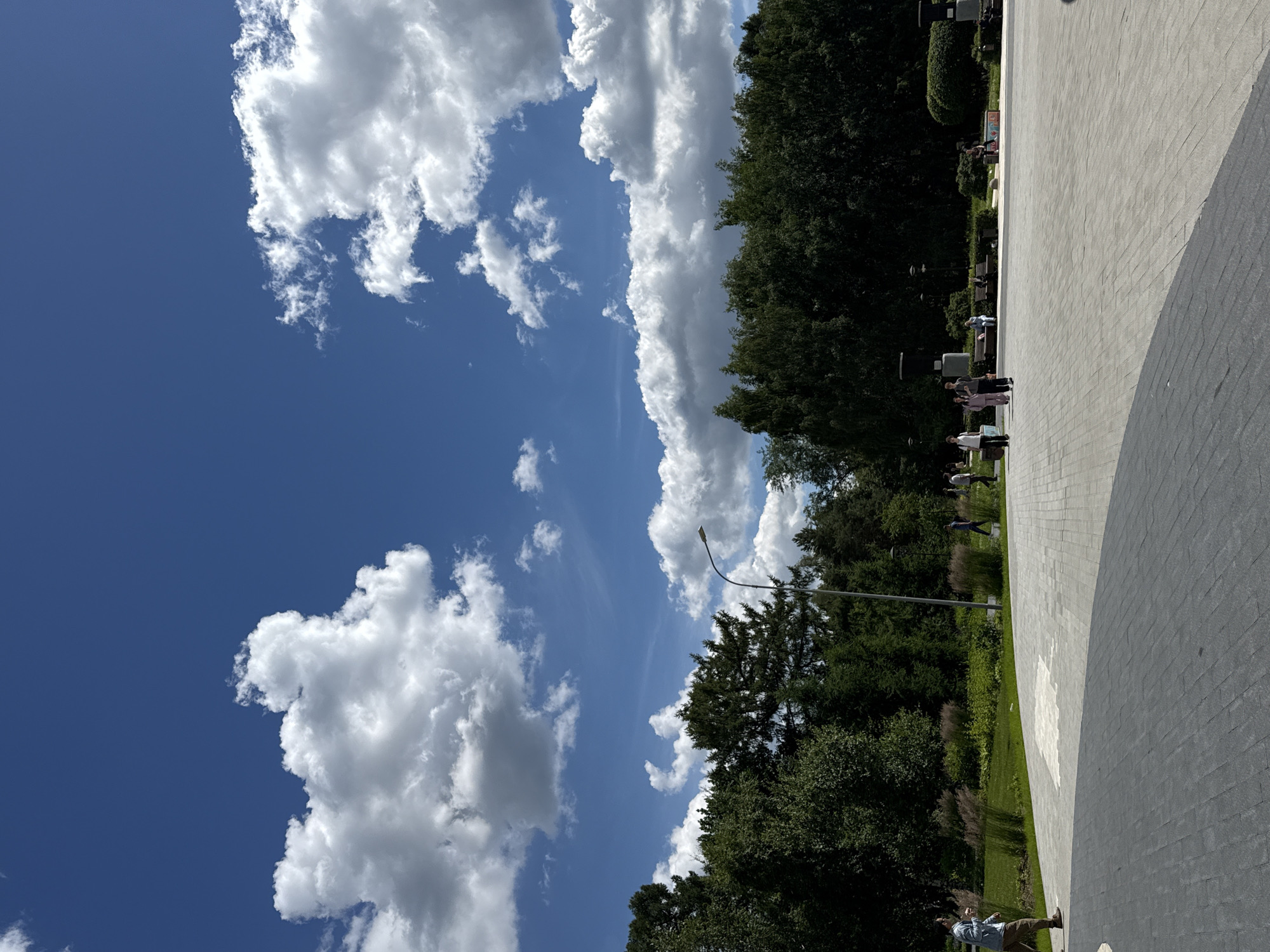 A bright, sunny day in Moscow, Russia, with a clear blue sky dotted with fluffy white clouds. The image is oriented vertically, as if the viewer is looking up towards the sky. To the right of the frame, a paved walkway curves into the distance. A line of trees runs along the edge of the walkway, creating a border between the paved area and a grassy park. Several people can be seen walking and standing on the walkway, enjoying the pleasant weather. Some are gathered in small groups, while others are walking alone. The trees are lush and green, suggesting it is summertime. A lamppost is visible among the trees. The overall scene evokes a sense of calm and relaxation, with people taking advantage of a beautiful day in a public park.