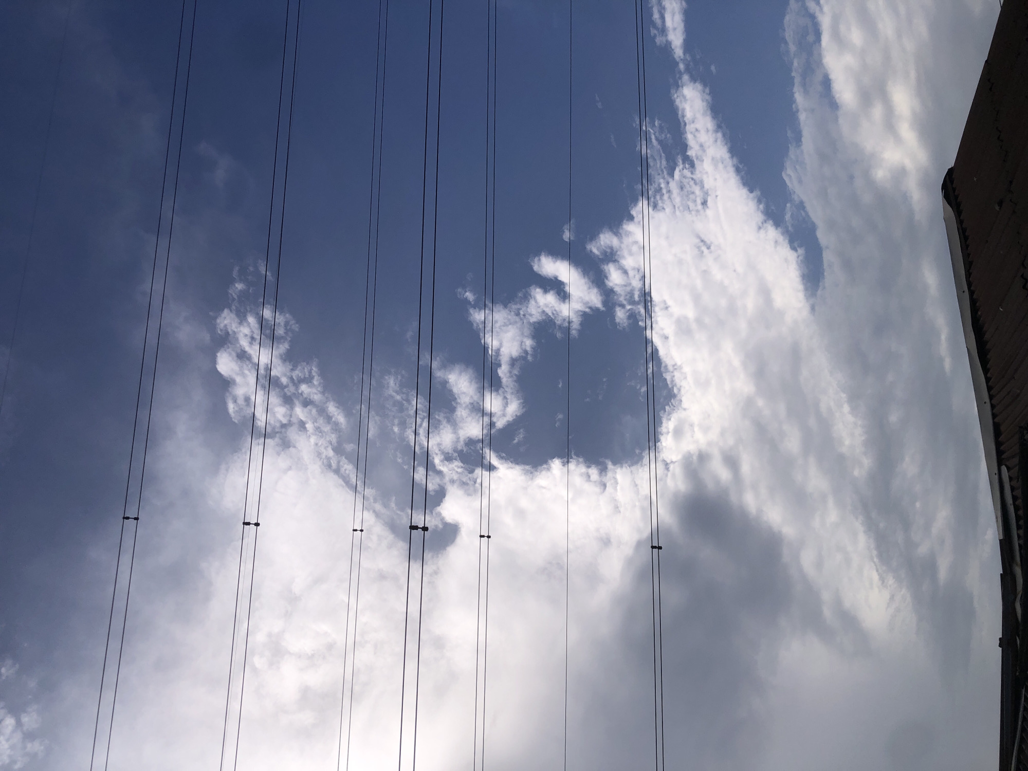 The image captures a cloudy sky above a corrugated iron roof in Ado, Nigeria. Several parallel electrical wires are strung across the frame, dividing the sky into horizontal bands. The clouds are a mix of white and gray, with patches of blue sky visible. The roof in the foreground is dark and appears aged, with some sections slightly bent or damaged. There are no people or animals visible in the frame.