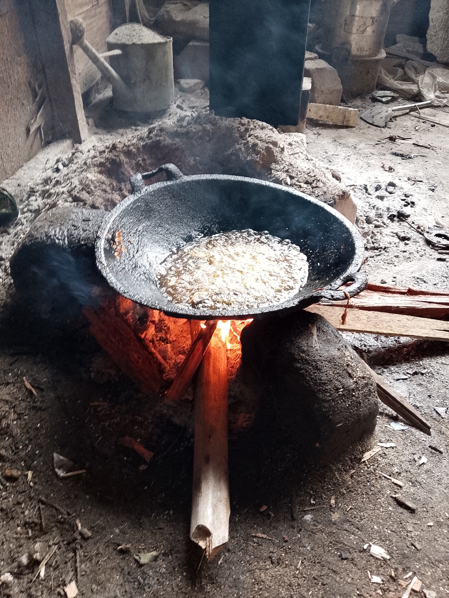 The image depicts an outdoor cooking scene, likely in a rural or semi-urban setting within Lhokseumawe, Indonesia. The focal point is a large, dark wok filled with bubbling oil, suspended over an open fire. The fire, fueled by logs, emits a vibrant orange glow and some smoke. The wok rests on two large, dark stones which serve as a makeshift stove. The ground around the fire pit is made of earth and scattered with debris, including small stones, wood chips, and ash. In the background, a rustic structure, possibly a shed or dwelling, is visible, with wooden beams and what appears to be a metal watering can covered in dust. The overall impression is one of resourcefulness and traditional cooking methods. The context of Lhokseumawe, Indonesia, suggests this scene is representative of everyday life in certain communities where such traditional cooking methods are still employed.