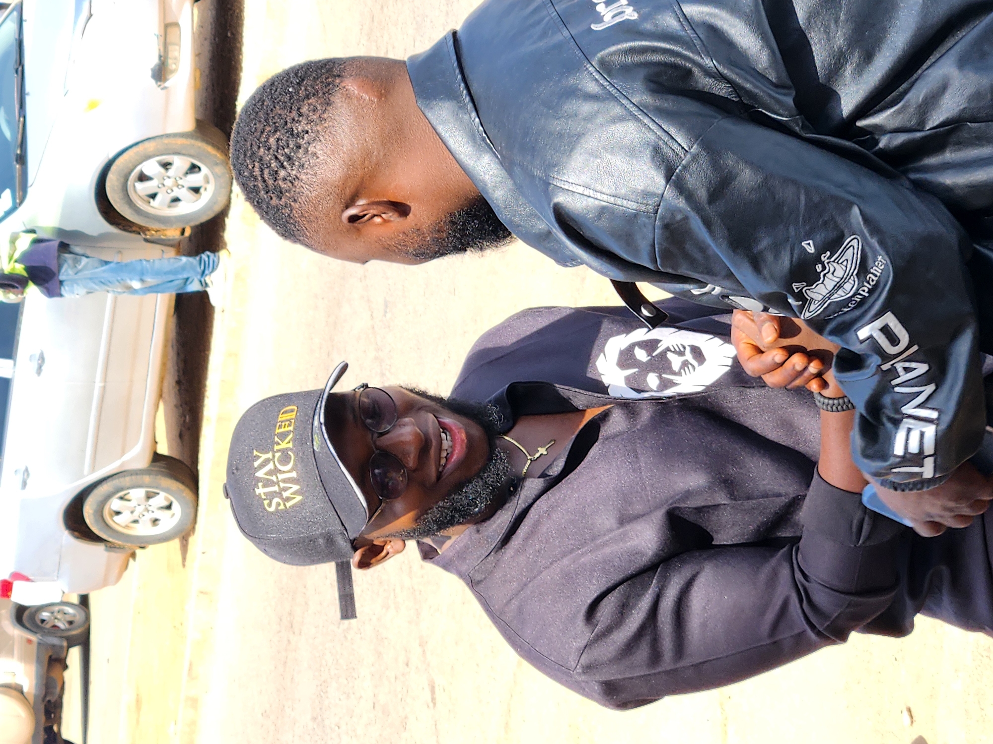 Two men are depicted interacting outdoors on a sunny day. In the foreground, a man with dark skin, a short beard, and sunglasses smiles broadly towards the viewer. He wears a black baseball cap with "STAY WICKED" emblazoned in yellow text, a black hoodie or jacket, and a thin gold chain around his neck. His left wrist has a black textured wristband.
The second man, positioned slightly behind and to the right, also has dark skin and a short beard. He is dressed in a black leather jacket. On his left sleeve, a white embroidered logo resembling a stylized planet or orb is visible, accompanied by partial text "en planet" and the more prominent "PLANET" written below it. His hands are clasped or interlocked with the hands of the first man, suggesting a handshake or friendly gesture.
The setting appears to be an open outdoor space with a light brown ground surface, possibly a dirt or paved area. In the background, two white cars are visible. One car is partially obscured behind the second man, while another is further back, with a person in a light blue shirt, jeans, and possibly a high-visibility vest standing near it. The strong sunlight indicates it is daytime, casting clear shadows. The overall impression is one of casual interaction and camaraderie between the two men.