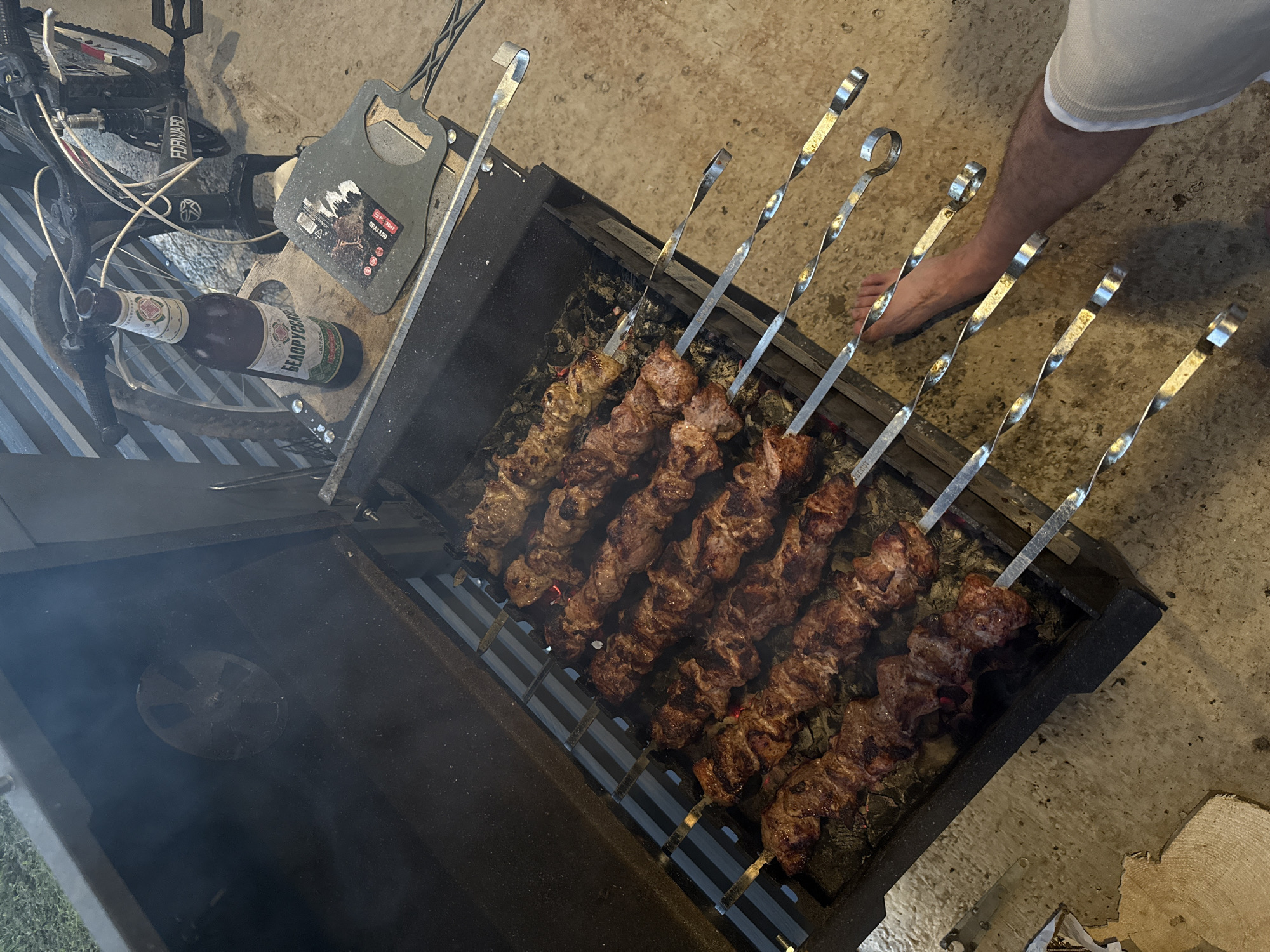 An overhead, slightly angled shot captures a close-up view of a barbecue grill laden with skewers of meat cooking over hot coals. The grill, with its lid partially open, is filled with multiple rows of these skewers, each holding chunks of seasoned meat. Smoke rises from the grill, indicating the cooking process is underway. To the right of the grill, a person's bare foot and lower leg are visible, suggesting they are standing close to the grill. In the background, a bicycle is partially visible, leaning against a structure with corrugated metal siding. A bottle of "BELORUSSKOE SVETLOE" beer rests on a wooden surface near the grill, along with a metal tool that appears to be a grill scraper or spatula. The ground is a concrete surface, and to the left, a cut log is visible. The scene appears to be outdoors, possibly in a backyard or outdoor recreational area in Podshivalovo, Russia.