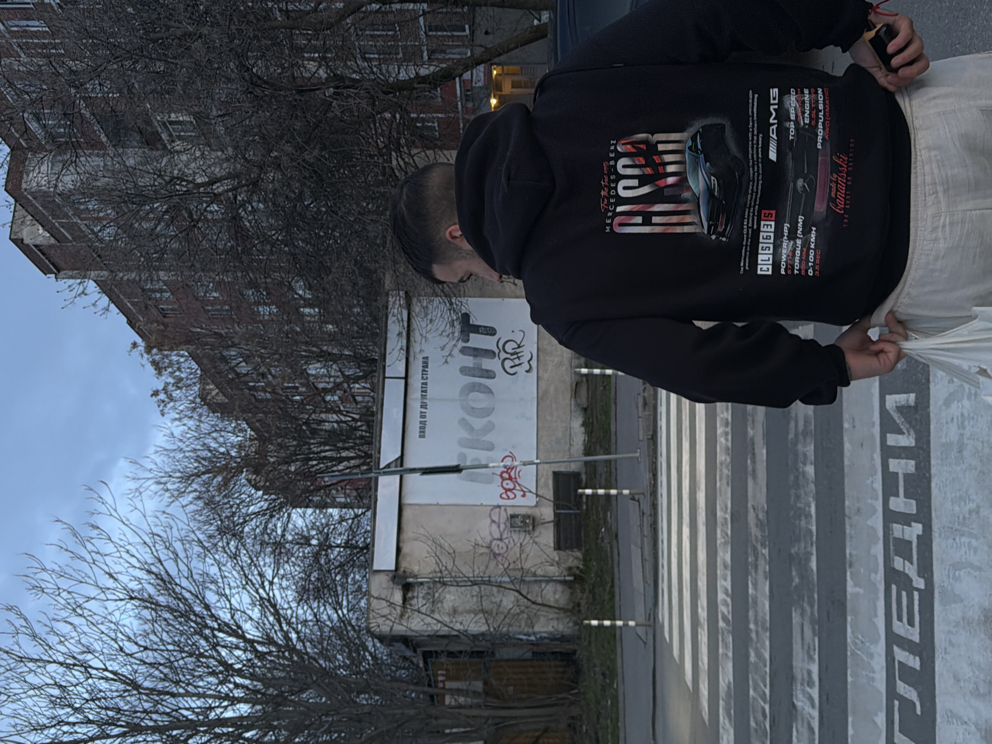 The image shows a person wearing a black hoodie with a car design on the back. They are standing on a crosswalk with the Bulgarian word "ГЛЕДНИ" (meaning "Look") painted on the ground. In the background, there is a dilapidated building with graffiti and signage. Based on the Cyrillic text on the signage and the style of the buildings, the image is likely taken in the city of Sofia, Bulgaria. The scene is in an urban environment with bare trees and buildings visible in the background.