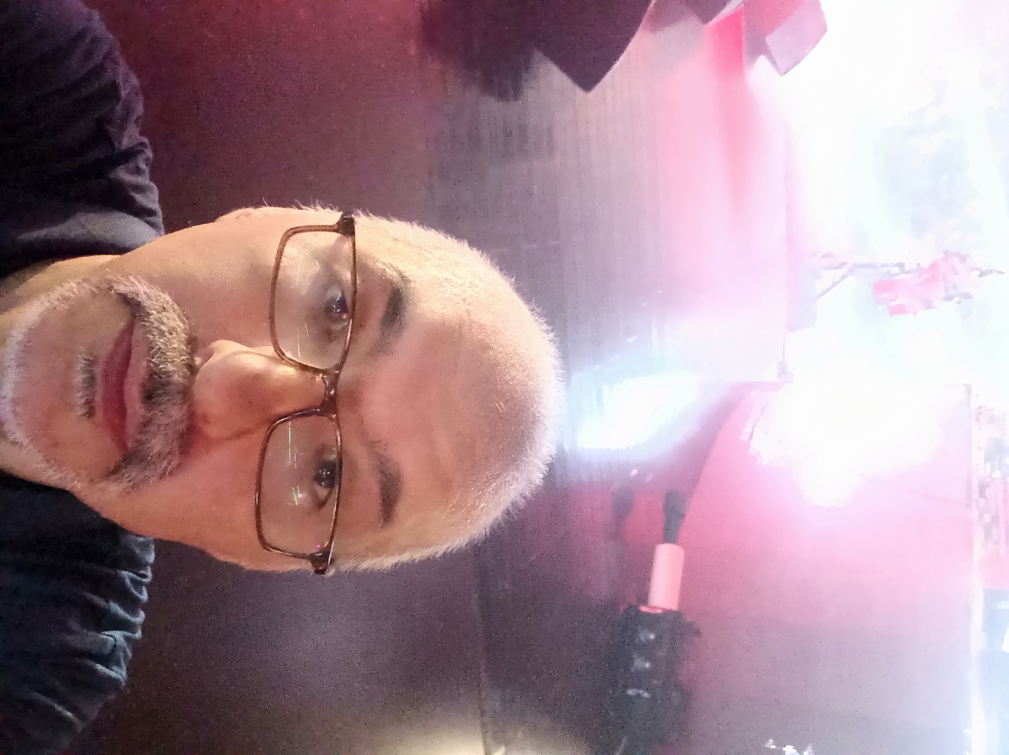 This is a selfie taken by a light-skinned man wearing glasses and a dark shirt. He has a mustache and a light goatee. The background is out of focus and appears to be a dimly lit indoor space with bright pink and white stage lighting visible. A microphone stand and part of a speaker can be seen in the background. The man is centered in the frame, with his face taking up most of the image. The image quality is grainy and has a strong pinkish hue from the lighting. There is no specific information within the image to suggest a location in Dushanbe, Tajikistan.