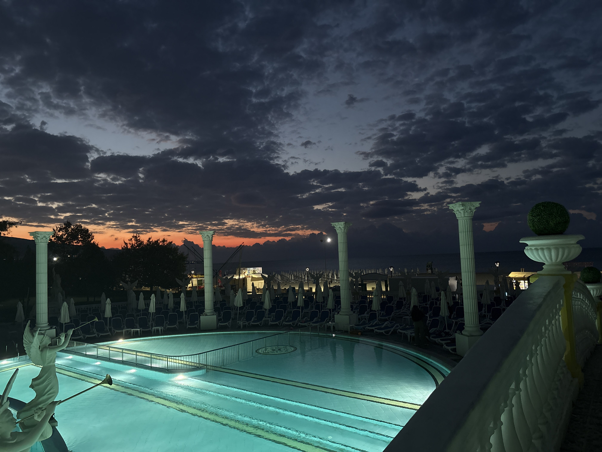 The image depicts an outdoor swimming pool area at dusk, with a dramatic sky filled with dark clouds, tinged with orange and red from the setting sun near the horizon. The pool itself is illuminated with bright turquoise lighting, reflecting the sky's colors. Surrounding the pool are rows of empty lounge chairs and umbrellas, suggesting a resort or hotel setting. Classical-style white columns and statues, one appearing to be an angel figure with a horn, are prominent architectural features. In the background, beyond the pool area, lights from buildings and possibly a beach are visible, with a dark body of water, likely the Black Sea, and a faint outline of hills or mountains in the distance. The location is identified as Nesebar, Bulgaria.