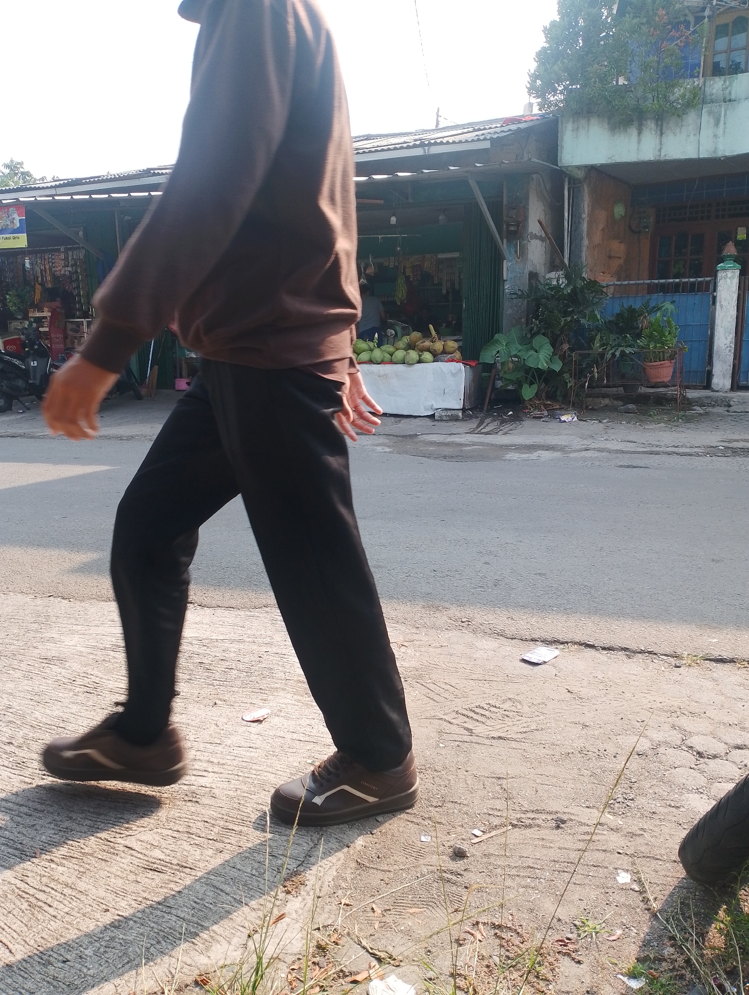 A person walks on a concrete sidewalk next to a paved road. The person is wearing a brown long-sleeved shirt and black pants. Their right hand is visible, extended backwards, with fingers slightly curled. They are wearing brown athletic shoes. In the background, there is a street stall selling coconuts. Behind the stall, there are buildings and trees. The scene appears to be in an urban or semi-urban area, possibly in Bogor, Indonesia, given the presence of coconuts and the architectural style. The lighting suggests it is daytime, with sunlight casting shadows.
