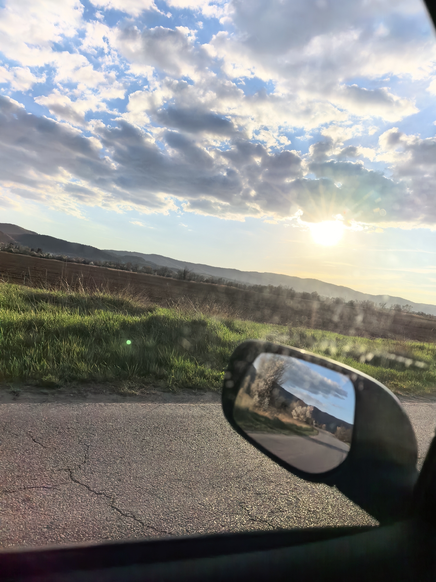 The image presents a serene rural landscape viewed from a vehicle during either sunrise or sunset, indicated by the low, bright sun creating a strong sunburst effect and lens flares in the upper right quadrant. The sky is a prominent feature, displaying a mix of clear blue patches and numerous fluffy white and grey cumulus clouds.
In the mid-ground, a range of rolling hills or distant mountains stretches across the horizon, partially obscured by a light haze. Below the mountains, the landscape consists of fields, some showing tilled brown earth and others covered in vibrant green grass. Sparse trees are scattered across the distant terrain and fields.
The immediate foreground reveals a section of an asphalt road with visible cracks, suggesting the vehicle's position on a well-traveled route. A significant element is the vehicle's side mirror, positioned in the lower right. This mirror reflects a portion of the scene behind the viewer, clearly showing a curving road, some light-colored trees (possibly in bloom), and a reflection of the sky with clouds.
The image captures the experience of traveling through a scenic countryside, emphasizing natural beauty and the interplay of light and clouds. The time of day, with the sun low on the horizon, lends a warm, tranquil atmosphere to the scene. No people are visible, nor is any text present in the image. The location appears to be a rural, possibly hilly or mountainous region, given the terrain.