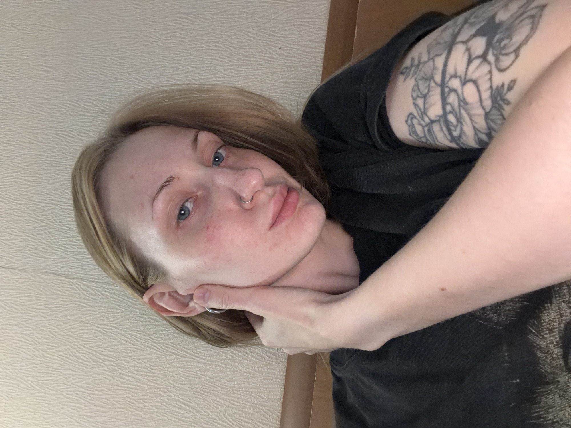 The media file shows a close-up, rotated, selfie-style shot of a young, fair-skinned person with blonde hair and blue eyes. They are wearing a black t-shirt and have a floral tattoo on their upper left arm. Their left hand is touching their cheek, near their ear. A nose ring is visible on their septum. The background is a textured, light-colored wall. The lighting appears to be indoor, with no strong shadows suggesting a specific time of day or weather condition. The person's expression is neutral to slightly pensive. The image is candid and personal in nature. There is no visible text in the media file.