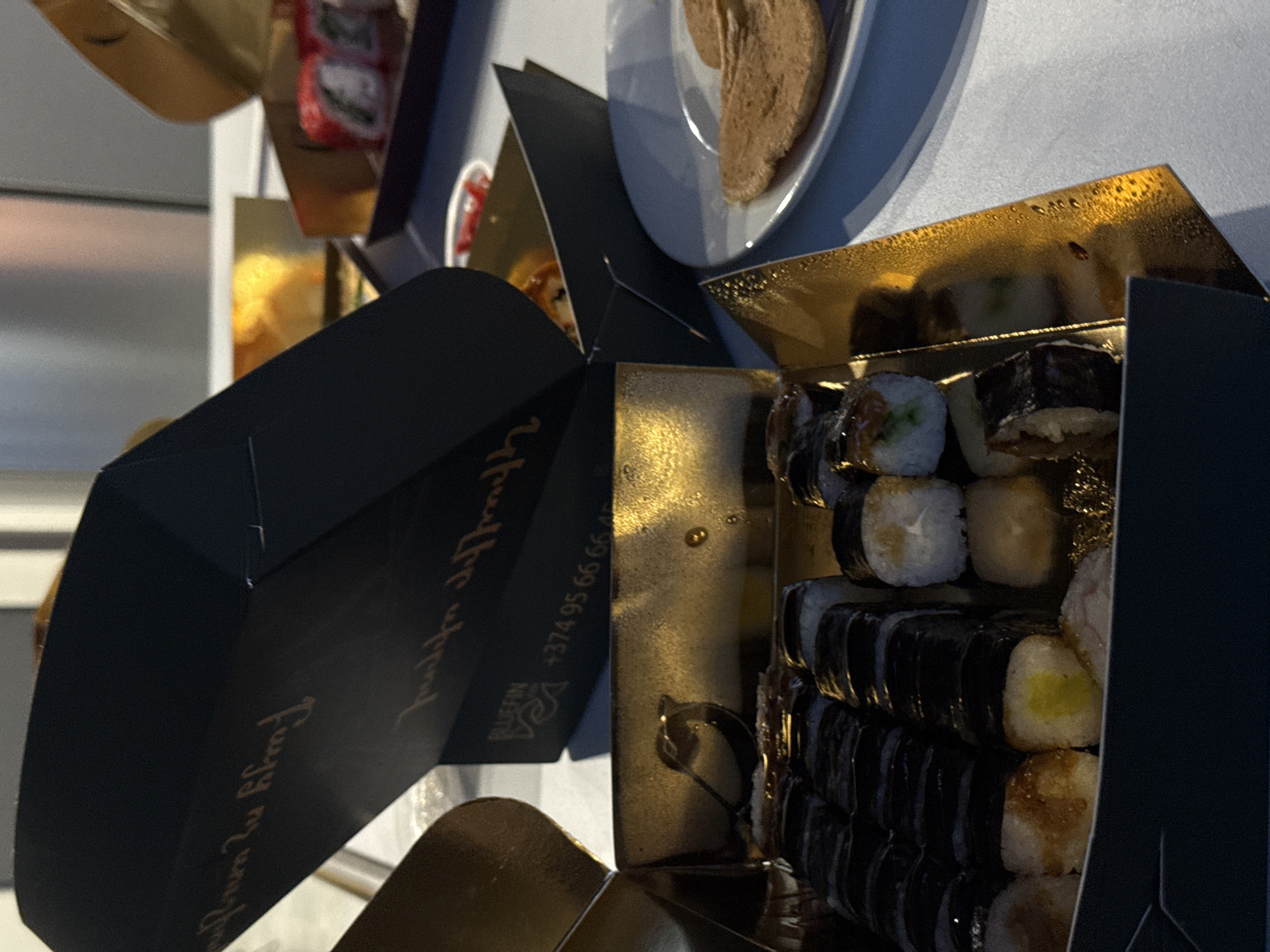 This close-up, angled photograph captures an open, multi-tiered black and gold sushi delivery box, likely in an indoor setting in Yerevan, Armenia. The box is filled with various types of sushi rolls, including maki and uramaki, arranged in multiple layers. Some rolls feature fillings such as cucumber, avocado, and what appears to be yellow pickled radish, while others show toppings or ingredients like salmon or a creamy sauce.
The interior of the box is a reflective, textured gold, showing subtle condensation or moisture, contributing to the impression of fresh or chilled food. Visible text on the black exterior includes the brand name "BLUFFIN" and an Armenian phone number: "+374 95 66 66". Another section of the packaging displays cursive gold lettering, deciphered as "Enjoy your meal".
In the soft-focus background, parts of other food items are discernible, including a white plate holding a brown, flat pastry or pancake-like item, and a small container with a red label. The overall scene suggests a meal delivery or catering event, with the focus on the organized and appealing presentation of the sushi. The lighting appears to be artificial, highlighting the textures and colors of the food and packaging. No individuals are clearly visible, and the image does not provide cues for the exact time of day or weather.