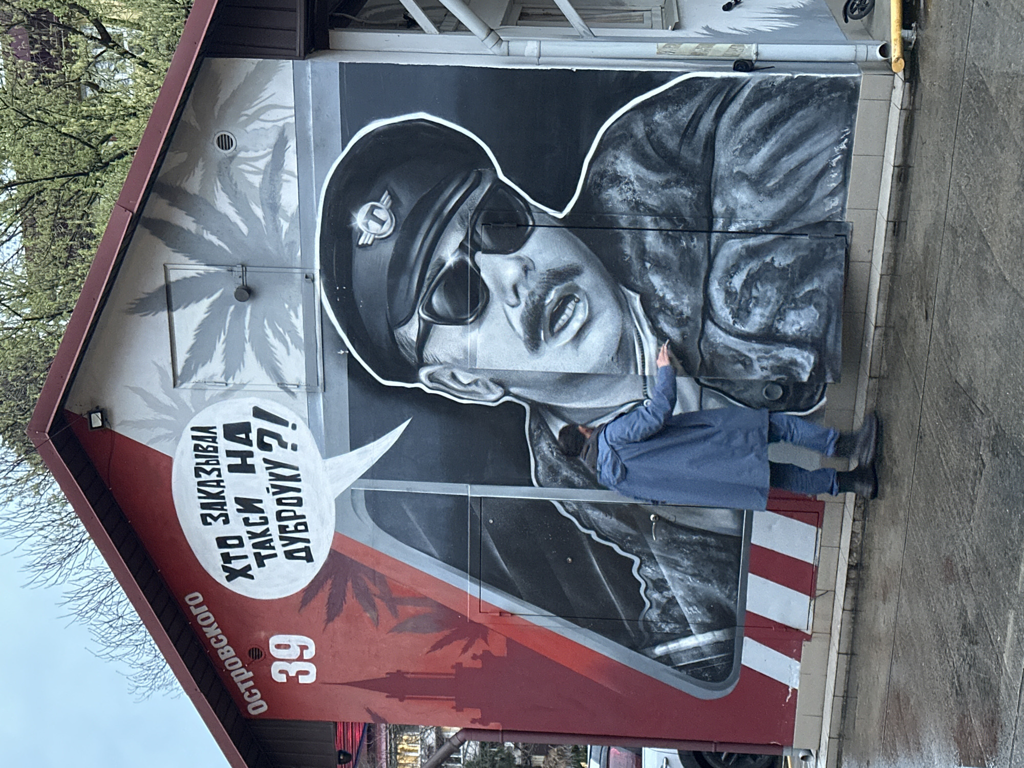 A street scene in Sochi, Russia, shows a red and gray building with a mural of a man wearing a beret and sunglasses. The mural incorporates a speech bubble with text in Russian, "Хто Заказывал Такси На Дубровуу??!", which translates to "Who Ordered A Taxi to Dubrovka?!". The building is numbered "39" and labeled "ОСТРОВоскОГО". A person in a blue coat, jeans, and black shoes is standing in front of the mural, with one hand raised towards the image. Red and white diagonal stripes adorn the lower section of the building. The surroundings include foliage and overcast sky.