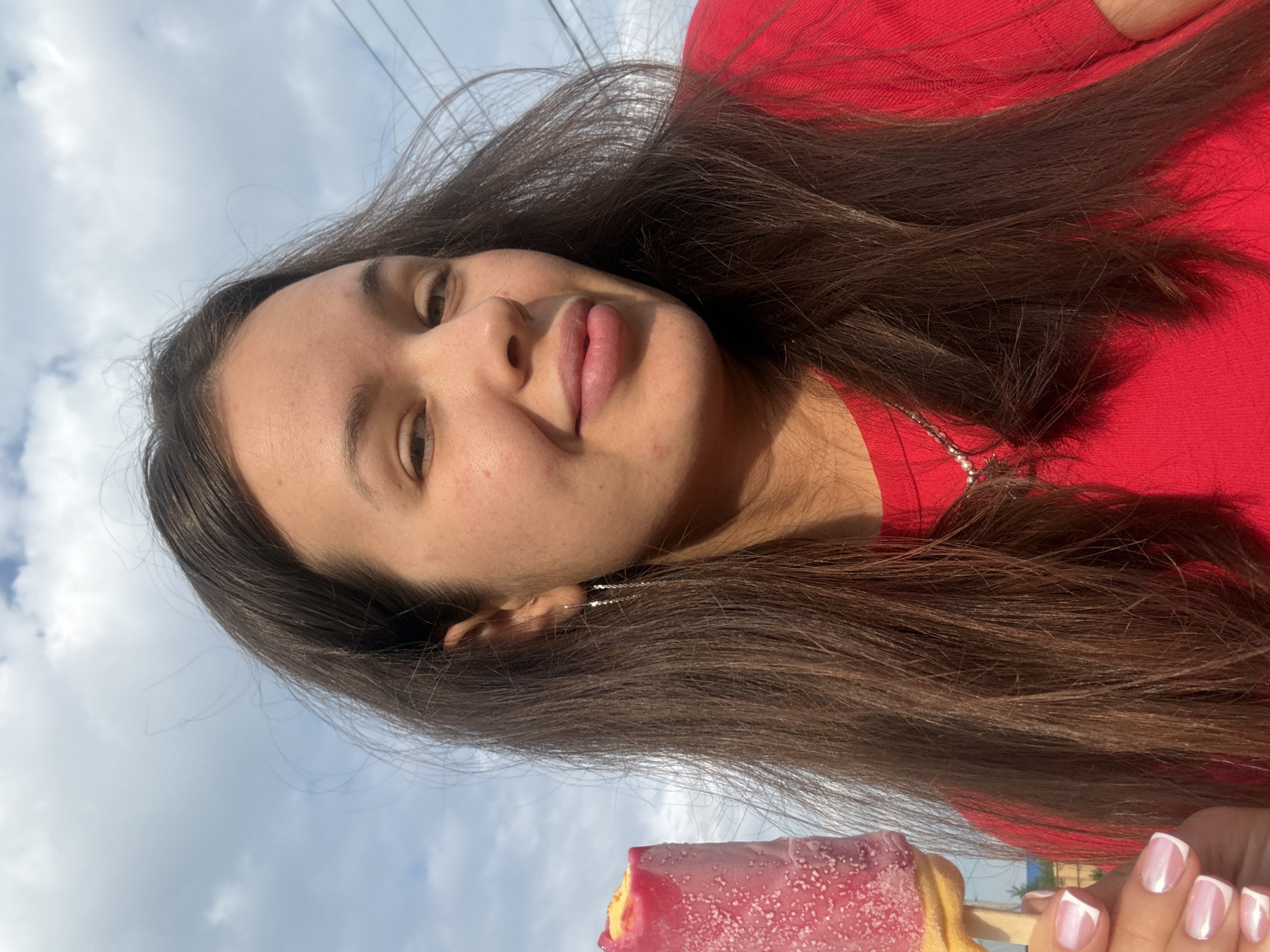 A young woman with long, dark brown hair is holding a pink popsicle and looking at the camera. She is wearing a red t-shirt and a silver necklace. The background shows a cloudy sky with power lines. She is outdoors, likely in an urban or suburban setting within Bely Yar, Russia, given the context provided. Her fingernails are painted white with a French manicure.