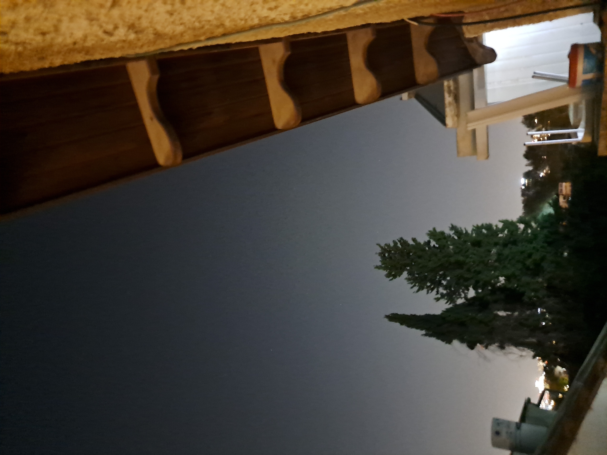 The image captures a night view from a balcony in Ramat HaSharon, Israel. The sky is dark with a few faint stars visible. In the foreground, a wooden ceiling with decorative supports extends from the right side of the frame. To the left, two tall, dark green trees stand prominently against the night sky, with faint city lights visible in the background. A grey cylindrical object, possibly a chimney or ventilation pipe, is partially visible on the left side of the frame, atop a building. The scene appears to be a residential area, with hints of buildings and lights suggesting an urban or suburban setting. The overall atmosphere is calm and quiet, typical of a nighttime scene.