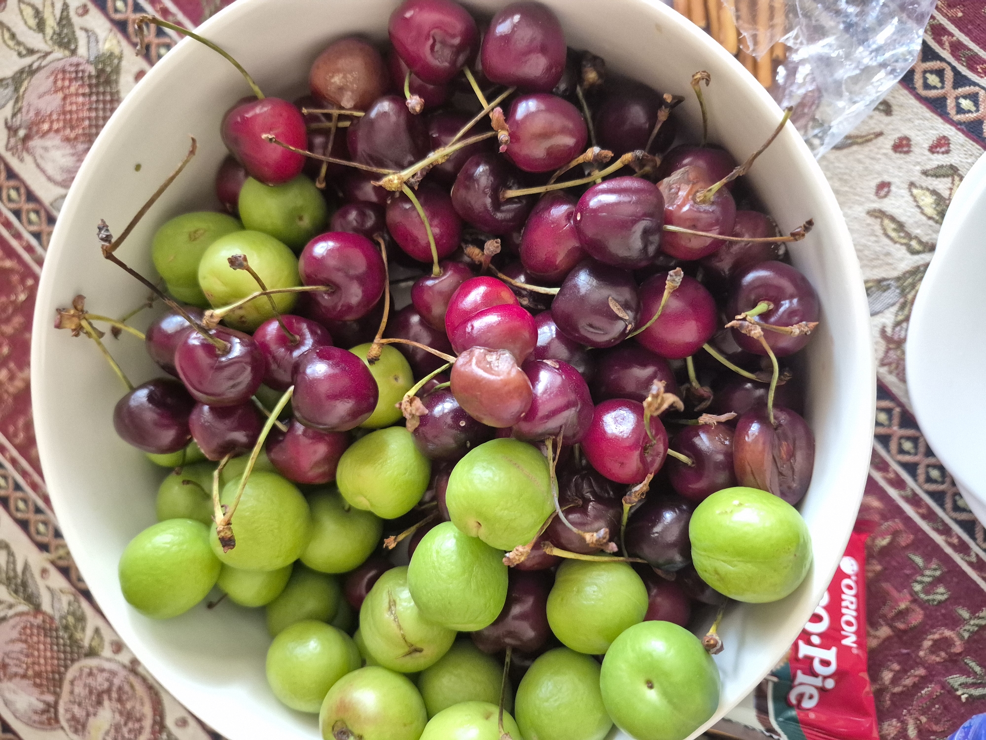 An overhead, close-up shot displays a white bowl brimming with a colorful mix of fresh fruits, resting on a patterned tablecloth. The bowl contains numerous dark red and deep purple cherries, many still attached to their stems, interspersed with an abundance of bright green, round plums. The fruits are densely packed, creating a striking visual contrast between the rich red hues and the vibrant green.
The tablecloth underneath features a traditional design with pomegranates and geometric patterns in shades of red, beige, and brown, suggestive of Armenian or regional aesthetics. In the upper right portion of the image, crumpled clear plastic wrap and the edge of another white dish are visible. A red food package is partially seen in the lower right corner, with the brand name "ORION" and the word "Pie" clearly legible.
The arrangement suggests the fruits are prepared for consumption, possibly as a snack or part of a meal in a casual setting. The natural, bright lighting indicates it is daytime. The green plums, given the Yerevan, Armenia context, are likely "goje" or "erik," a common sour plum consumed in the region. No individuals are present in the image.