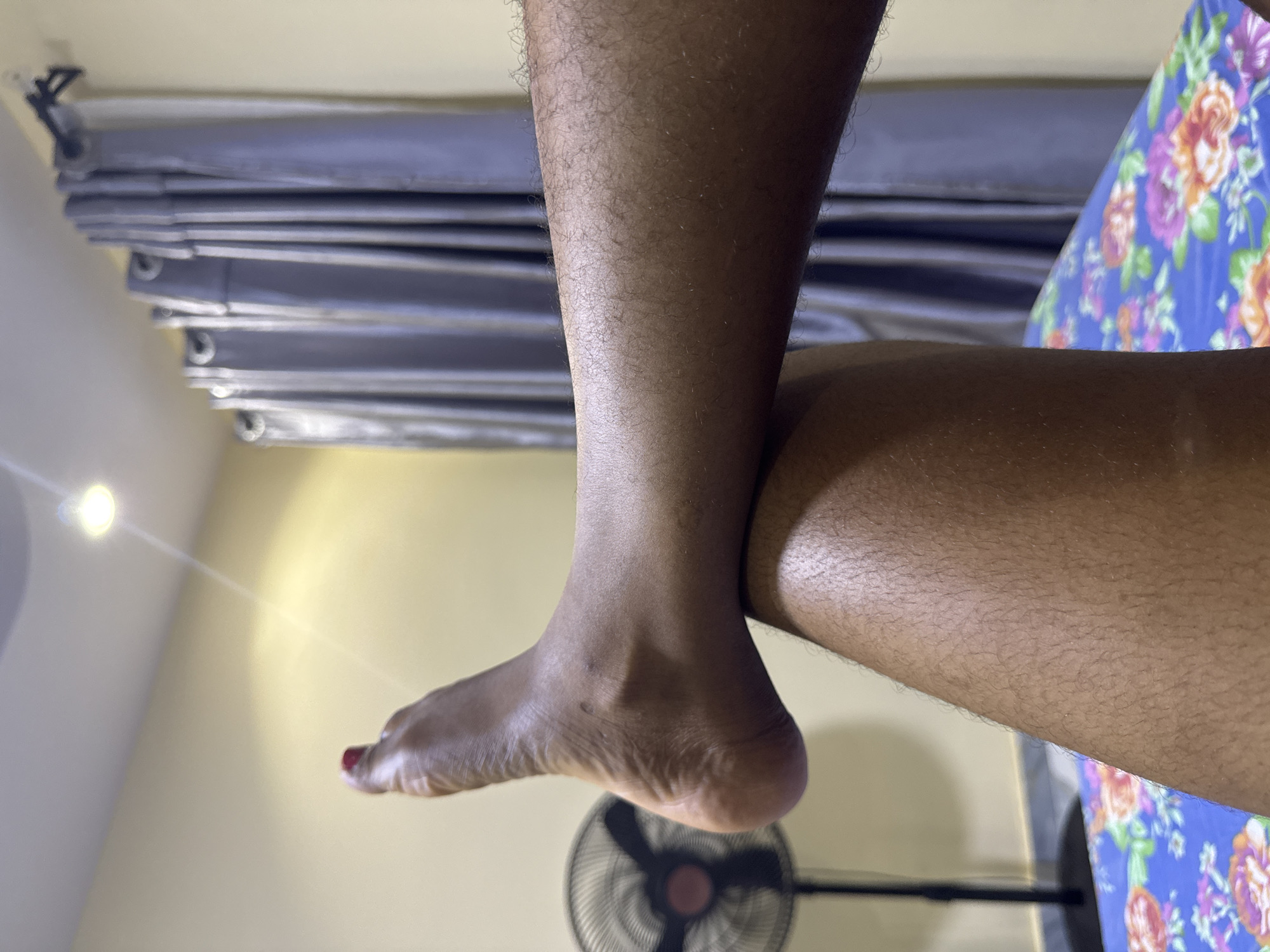 The image depicts a close-up view of a person's legs and feet. The feet are crossed, with one foot resting on the other leg. The skin is dark and appears to have some hair. The toenails on the visible foot are painted red. In the background, a black oscillating fan is partially visible, along with a light-colored wall, grey curtains, and a portion of a bed with a floral patterned sheet. The lighting suggests an indoor setting, possibly a bedroom. The scene is framed from a low angle, focusing on the lower extremities of the individual. The overall impression is intimate and casual.