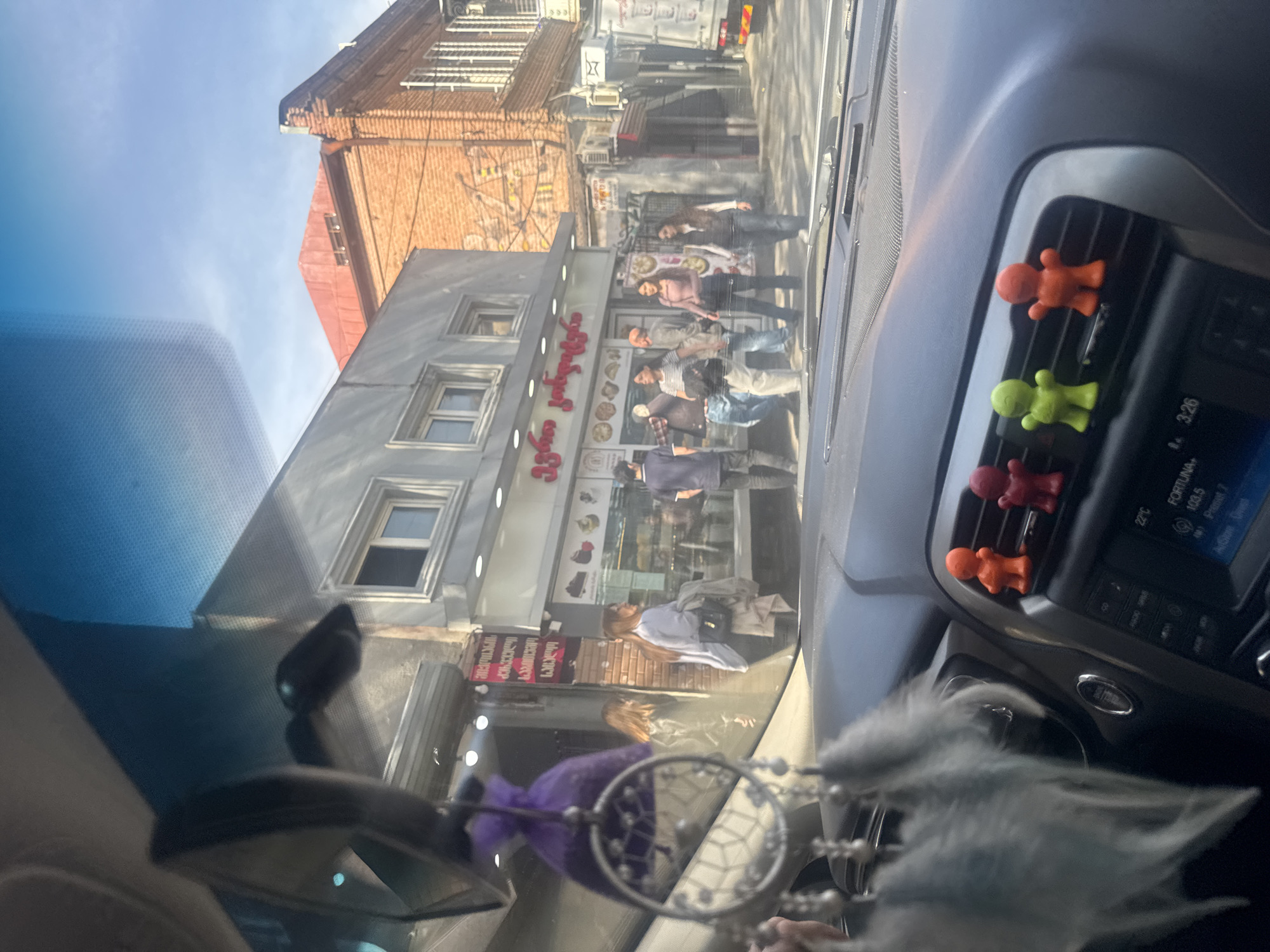 The image, taken from inside a car in Tbilisi, Georgia, shows a street scene reflected in the windshield. Pedestrians are visible on the sidewalk in front of a building. The building has Georgian script signage, likely indicating a business or establishment. The car's dashboard is in the foreground, featuring air vents adorned with colorful human-shaped decorations. A dreamcatcher-style ornament hangs from the rearview mirror. In the upper portion of the reflection, the street continues with buildings and a vehicle, possibly a truck or van, can be seen further down the road. The sky is visible in the upper left of the reflection.