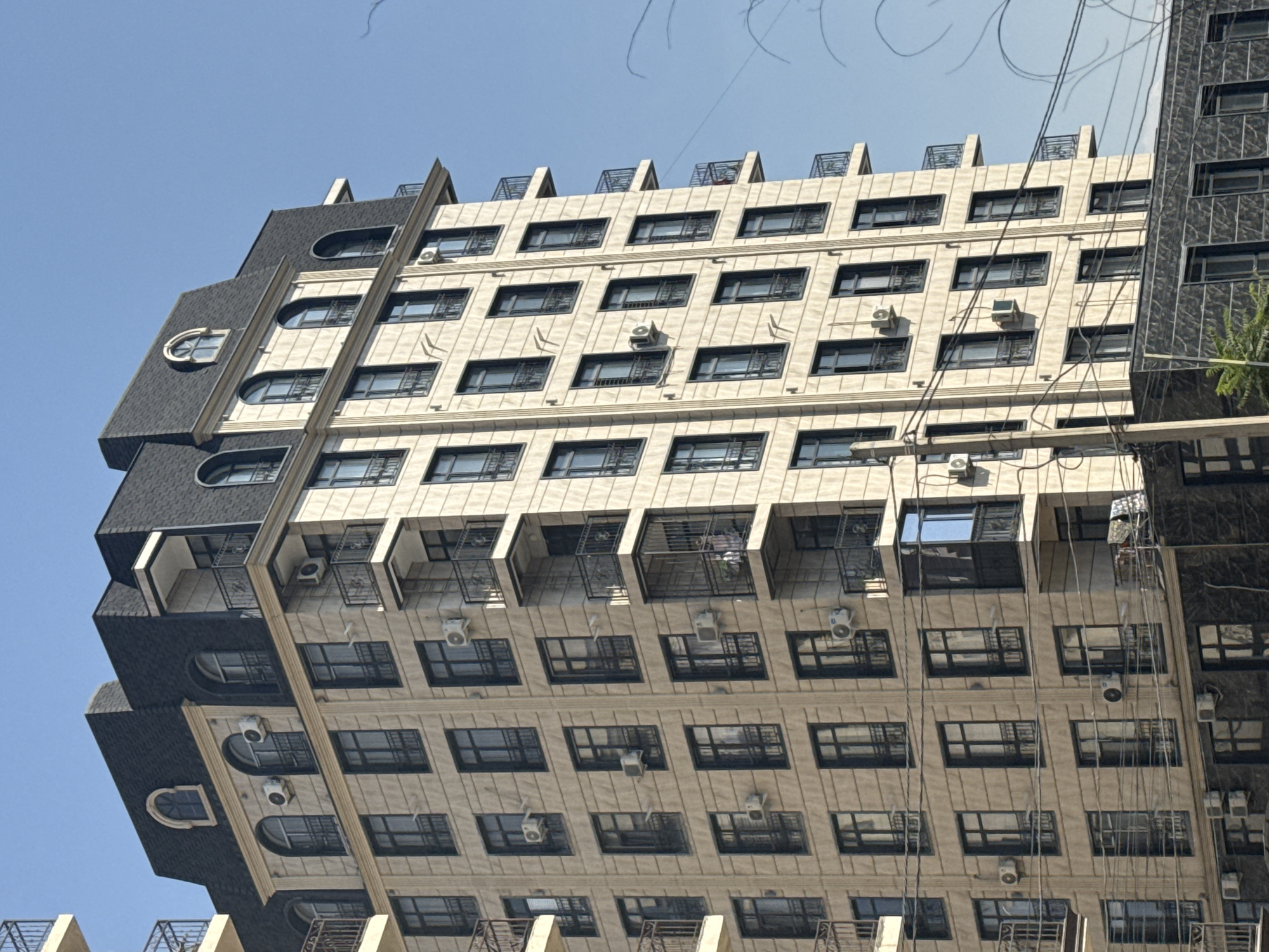 This image captures a close-up, slightly angled exterior view of a tall, modern apartment building in Bishkek City, Kyrgyzstan, under a clear blue sky. The building's facade is a light, textured stone, with dark-framed windows and numerous balconies. Many of the windows appear to have dark shutters or grates, and several window air conditioning units are visible. The building has a dark, mansard-style roof with arched dormer windows. In the foreground, power lines crisscross the view, obscuring some of the lower floors of the building. To the right, a portion of another, darker-colored building is visible, with similarly dark-framed windows. The scene is devoid of any visible people or activity, presenting a static architectural study.