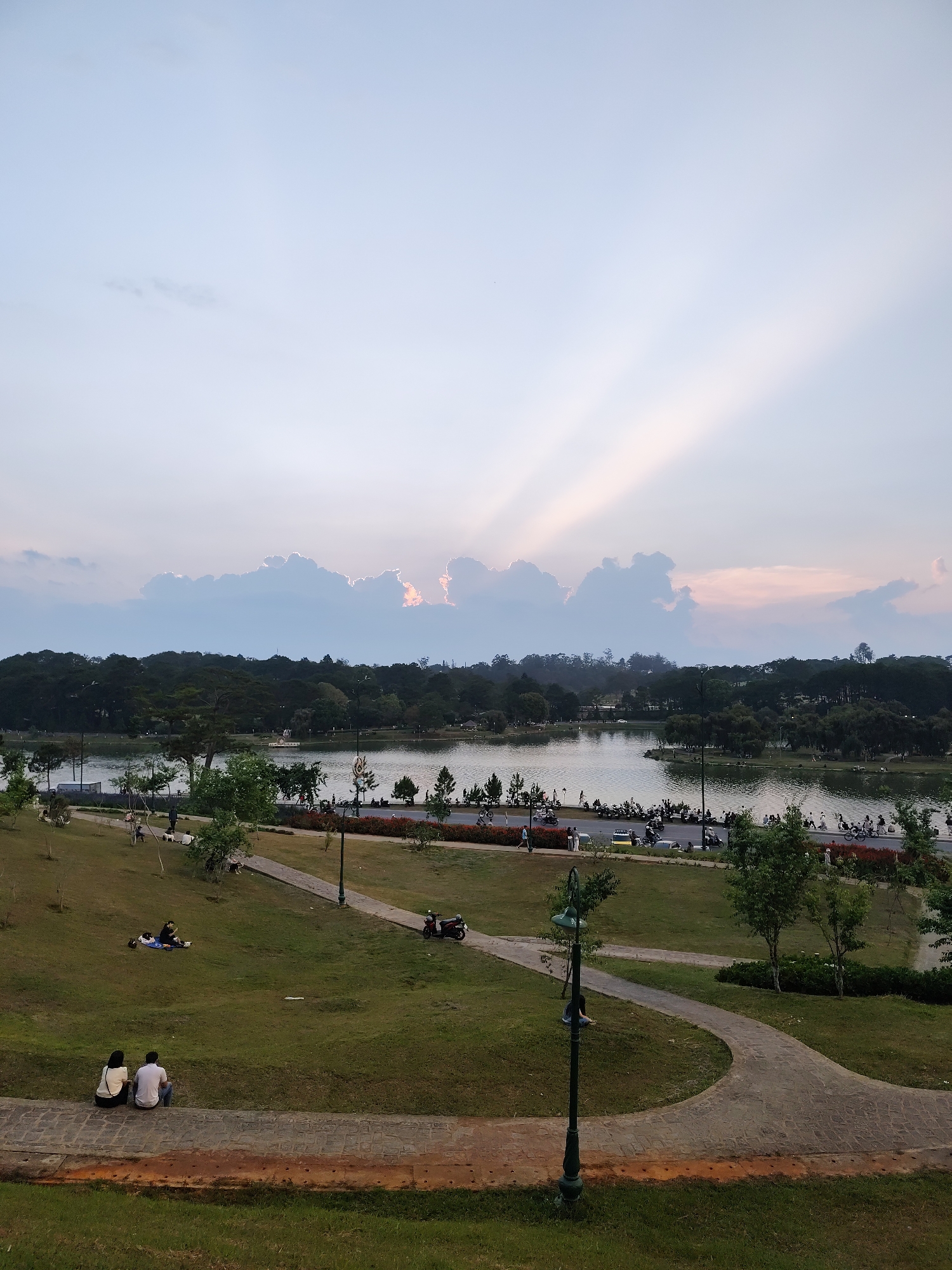 The photo depicts a tranquil scene at Xuan Huong Lake in Da Lat, Vietnam. The foreground features a grassy slope with brick-paved walkways. A couple sits on the path's edge, while another group relaxes on the grass. In the midground, the calm lake reflects the surrounding trees and sky. A road runs along the lake's edge with people and vehicles. In the background, a tree-lined landscape meets the horizon, capped by a cloudy sky with visible sun rays.