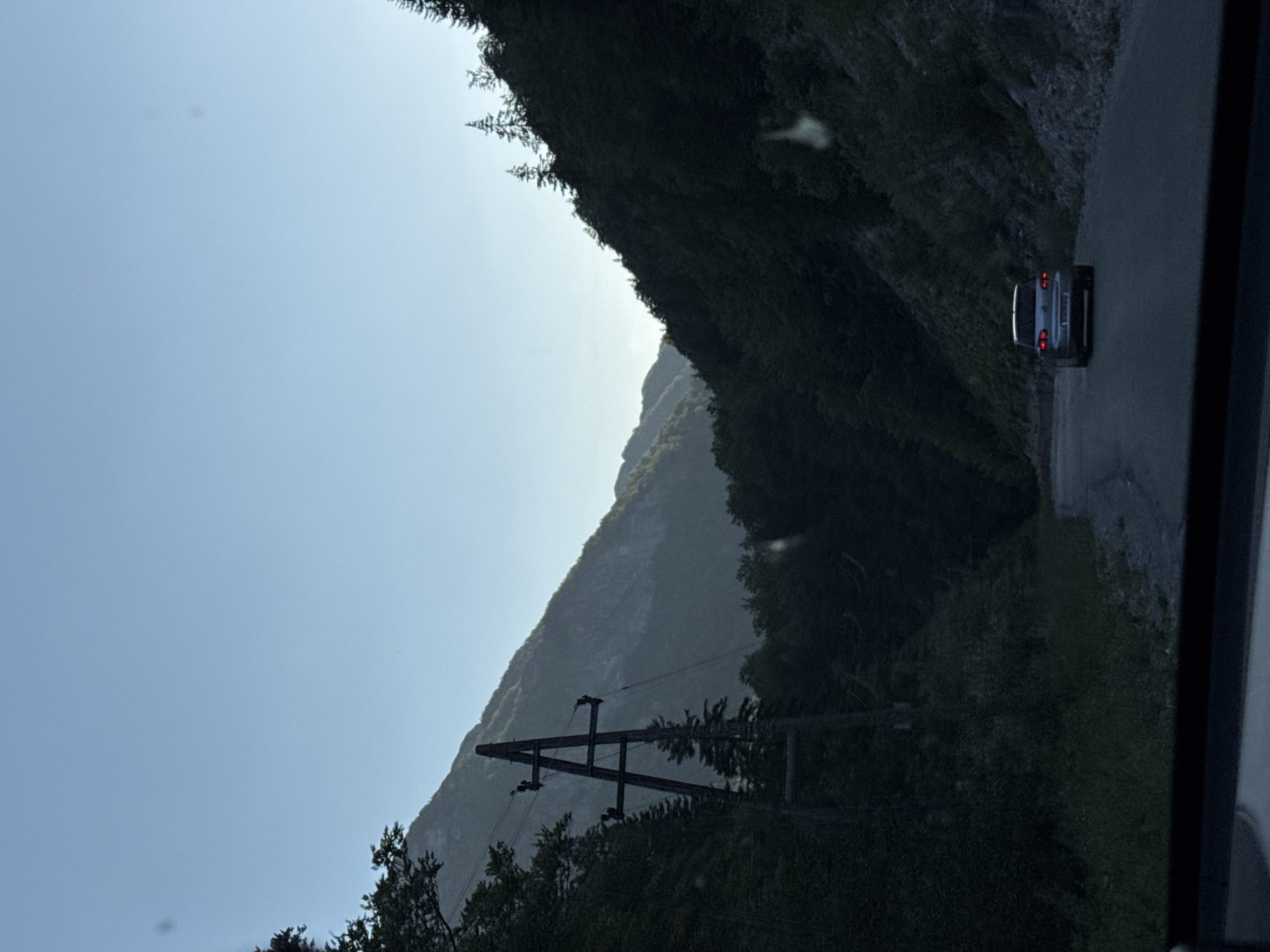 The image shows a silver car driving down a winding road through a forested mountain pass. The sky is clear and blue. A power line tower stands on the left side of the road, with trees lining the mountainside. The car is the primary subject, viewed from behind as it moves away from the camera. The environment is natural and mountainous, with dense vegetation covering the slopes. The scene appears to be captured from inside another vehicle, with the lower edge of the frame showing a dark dashboard. The lighting suggests it is either early morning or late afternoon.