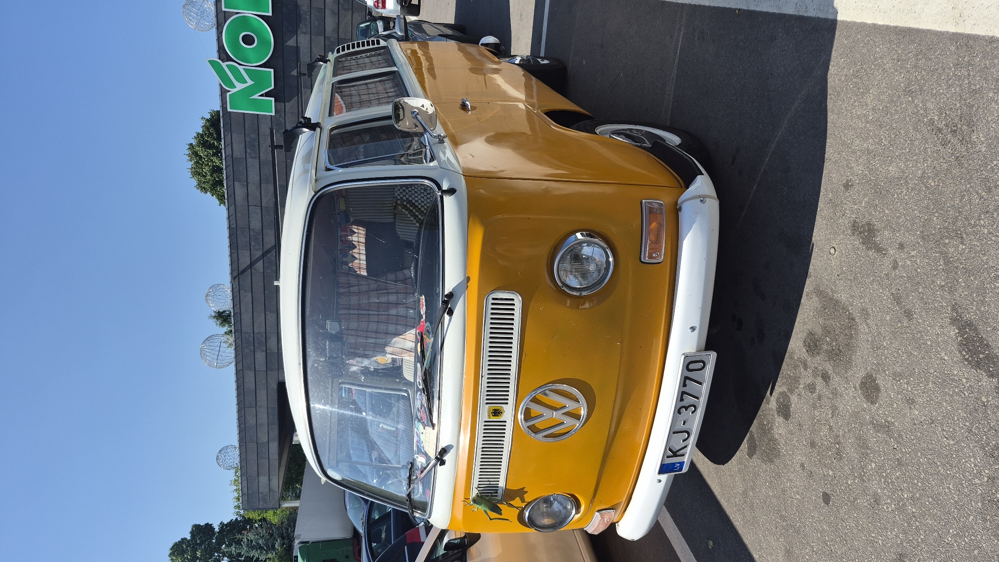 A vintage yellow and white Volkswagen Type 2 van, with license plate "LV KJ-3770", is parked in a lot on a sunny day. A green plastic grasshopper figurine is attached to the front of the van, near the left headlight. The van has a white roof rack and its windows are partially covered by checkered curtains. In the background, a building with a dark facade is visible, featuring large green lettering that appears to spell out "NOL" or a similar word. The sky is clear and blue. The parking lot surface is asphalt with visible white painted lines. The setting is likely a commercial area or parking lot in Zarasai, Lithuania, given the license plate format.