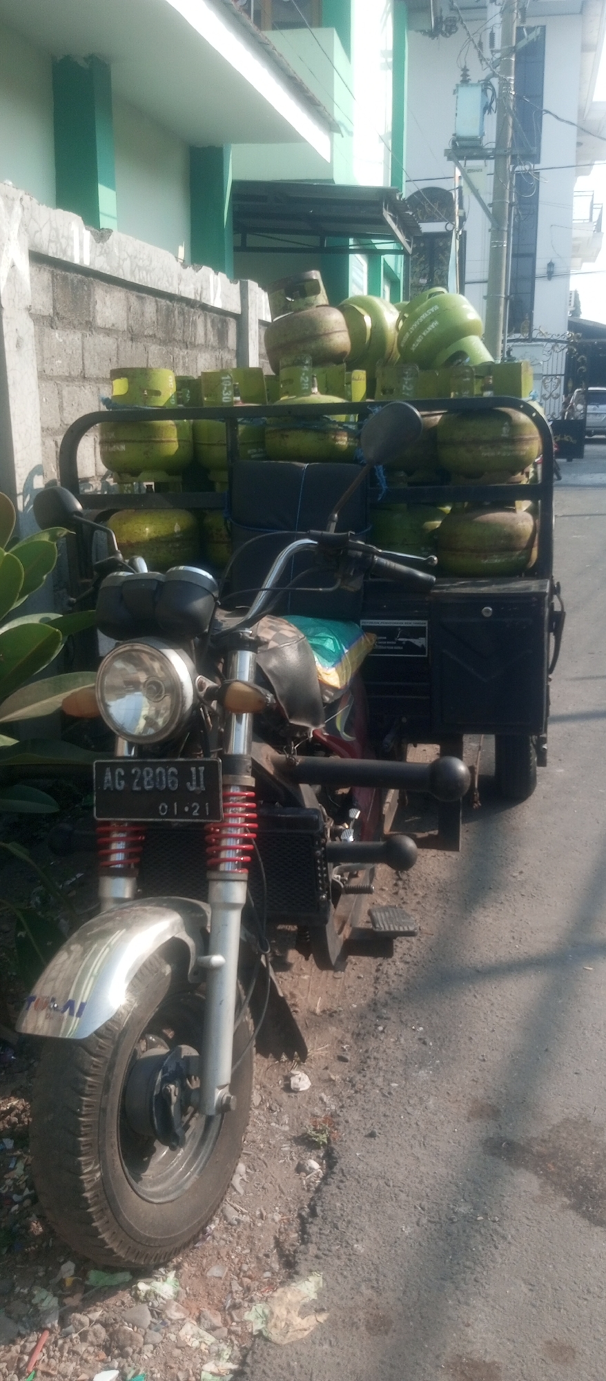 The image depicts a three-wheeled motorcycle, a type of vehicle commonly known as a "roda tiga" in Indonesia, parked on a roadside in Kediri City, Indonesia. The motorcycle is laden with numerous green and yellow LPG (liquefied petroleum gas) cylinders stacked on a black metal rack behind the driver's seat. The motorcycle itself is front-heavy, with a prominent round headlight and a black license plate in front, displaying the text "AG 2806 JI" and "01·21". The front wheel is silver and chrome, with red coil springs visible on the suspension. The surrounding environment includes a gray brick wall to the left, a light-colored building with green accents in the background, and a paved road with scattered debris. The scene is illuminated by bright sunlight, casting shadows on the ground. There are no people visible in the image.