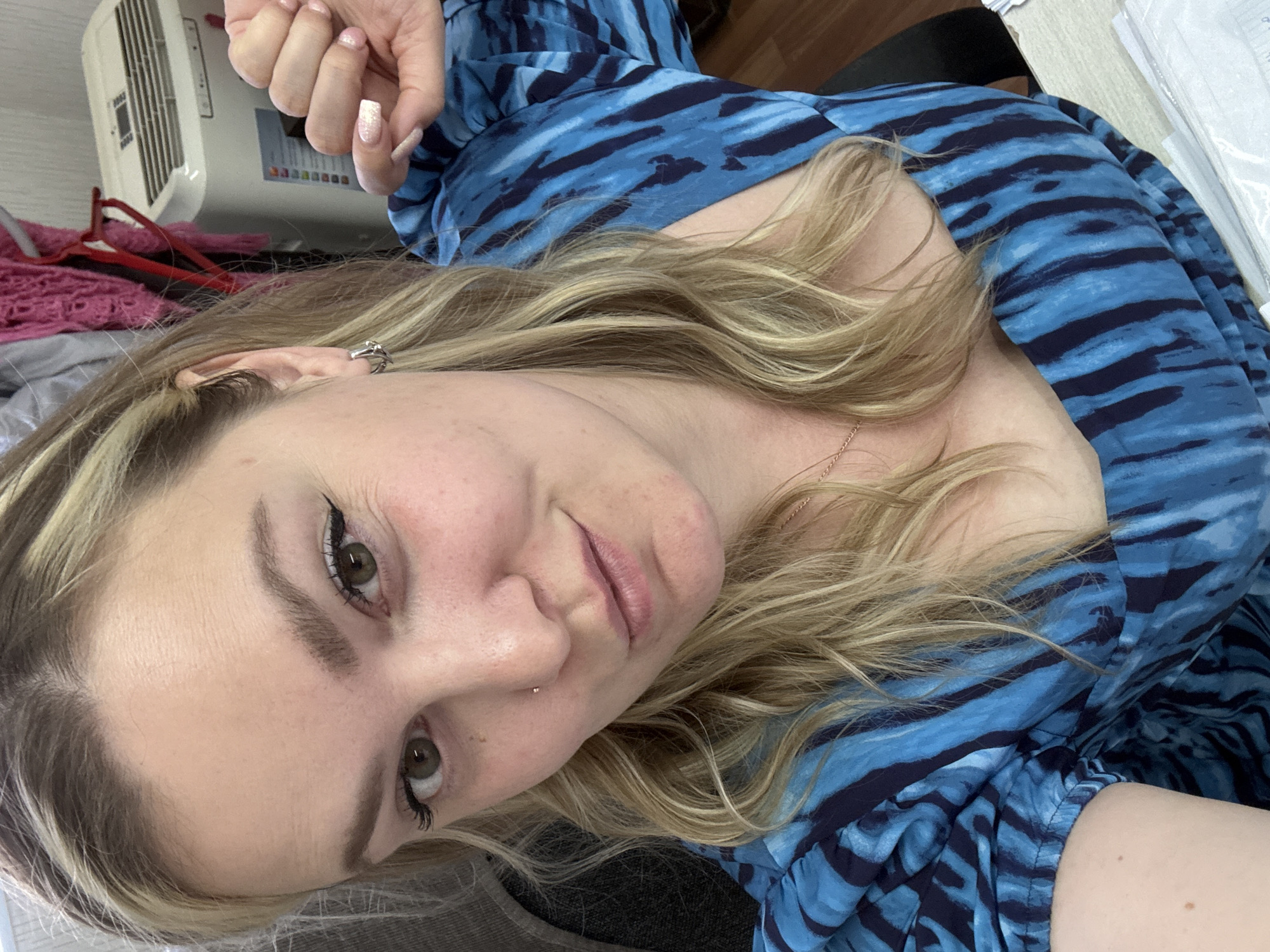 A close-up selfie of a blonde woman wearing a blue patterned dress. She has light skin, green eyes, and is wearing eyeliner. A small nose ring is visible. A portion of her hand with long, manicured nails is in the upper left corner. In the background are a white air conditioner and some indistinct items in pink and red. The location is unspecified, and there is no indication that this image was taken in Noviy, Russia.