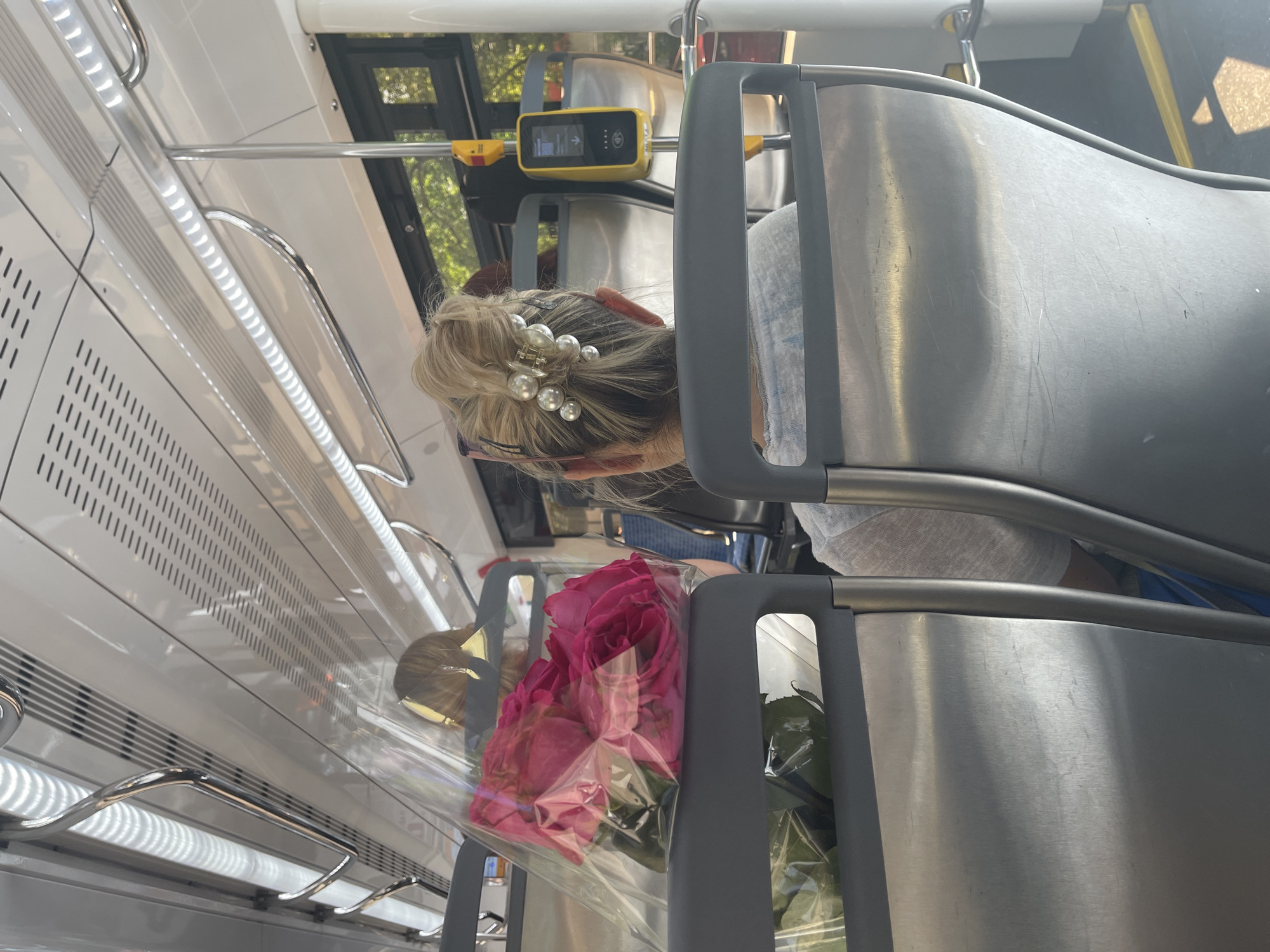 The image captures the interior of what appears to be a bus or tram in an unknown city. A blonde woman with a decorative hair clip is seated, and a bouquet of pink roses wrapped in clear plastic is placed beside her, secured by the seat. Overhead, a yellow transit payment device is visible. The interior features stainless steel panels and handrails, and rows of air vents. The scene conveys a sense of urban transit, with the woman and flowers perhaps representing a recent event or gift.
