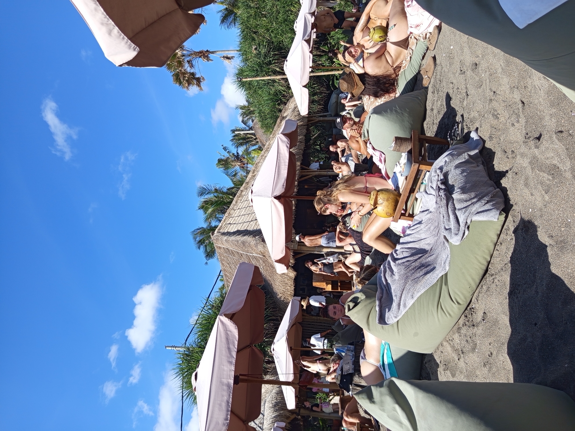 The scene is an outdoor beach setting in North Kuta, Indonesia, during daytime. A row of thatched-roof structures provides shade, under which people are lounging on cushions and beach chairs. Beach umbrellas are present, casting shadows on the dark sand. The sky is a clear blue with scattered clouds. Individuals are reclining, some holding drinks, and appear to be relaxing and sunbathing. The area is populated with a mix of people enjoying the beach environment.