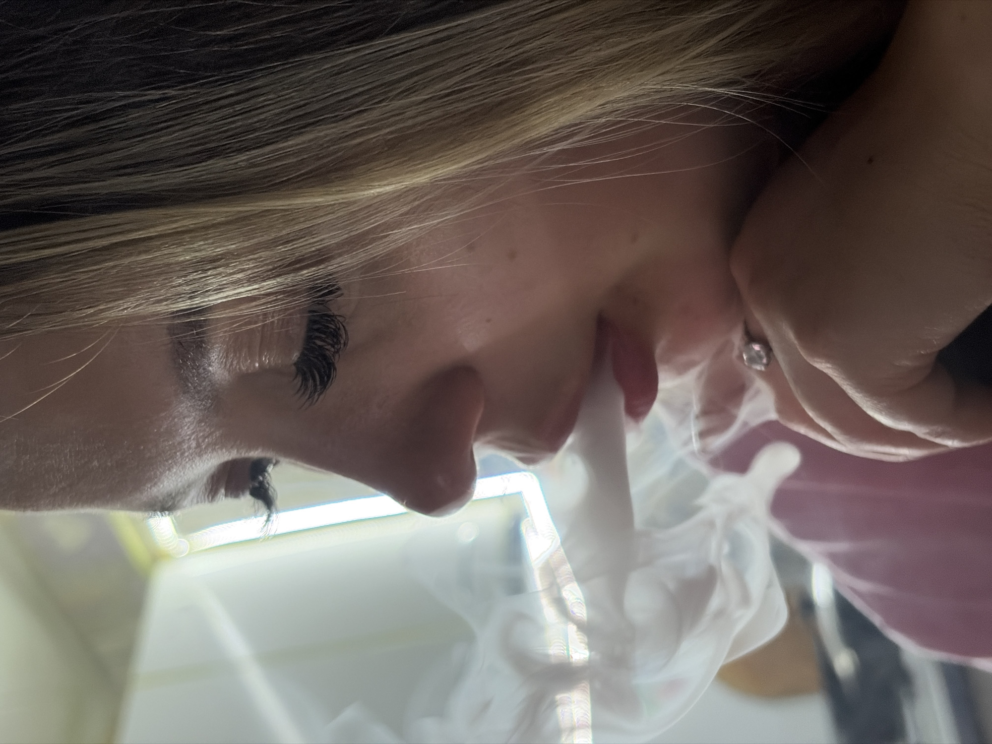 A close-up, angled view shows a person exhaling a thick cloud of white vapor. The individual has fair skin, dark eyelashes, and light brown hair. Part of a hand wearing a ring is visible near the person's face. The setting includes a reflective surface, potentially a table or countertop, and some indistinct objects. There is no information to suggest the location is in Port-Katon, Russia.