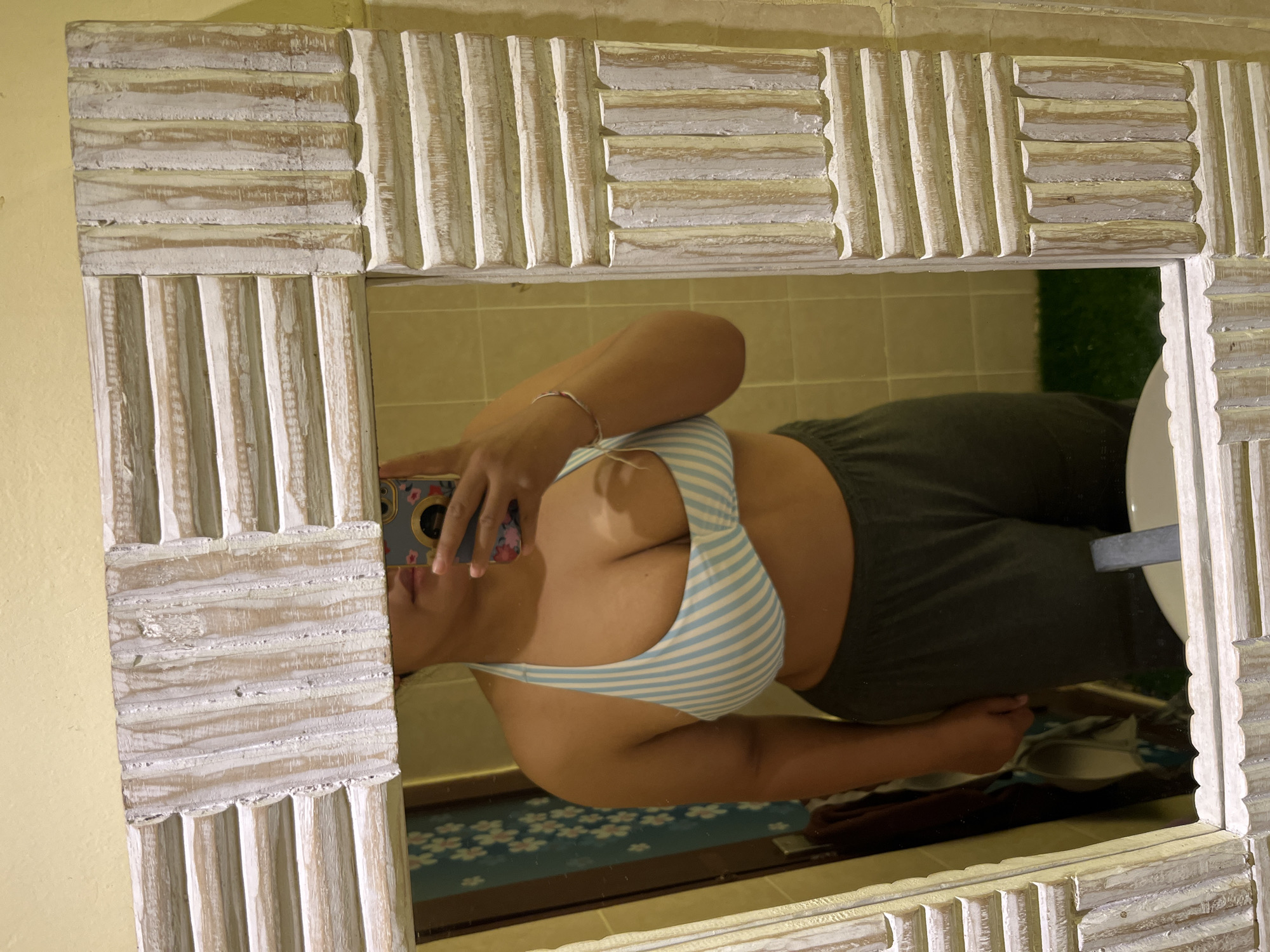 A person is standing in front of a mirror, taking a selfie. They are wearing a blue and white striped bikini top and dark grey sweatpants. The mirror has a distressed wooden frame with horizontal and vertical carved details. The person's hand is visible holding a smartphone. The background of the mirror reflects a tiled wall, a curtain with a floral pattern, and a glimpse of what appears to be a bathroom sink and faucet. The overall impression is a casual, indoor scene, likely a home or hotel bathroom in Kuta, Indonesia, given the context of the request.