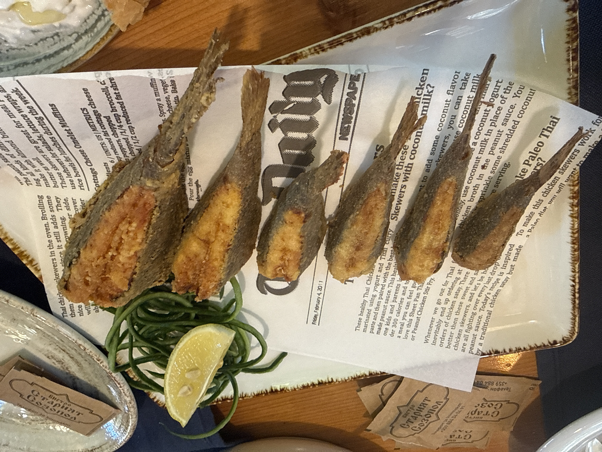 The image shows a close-up view of a plate of fried fish and skewers, served on top of newspaper clippings. The food is arranged on a white plate with a decorative rim, and a wedge of lemon and a pile of green vegetables are placed alongside the fish. Two small packets, with Cyrillic writing that includes "Бистро Старият Созопол," are also visible on the plate, suggesting the location is a bistro in Sozopol, Bulgaria. The newspaper clippings contain text and images, some of which are recipes and articles related to food, with one date visible as "Friday, February 4, 2011." The overall scene appears to be a meal being presented, likely in a casual dining setting. The lighting suggests it could be daytime, and there are no indications of weather conditions. The focus is on the food presentation, with the newspaper providing a rustic and thematic backdrop.