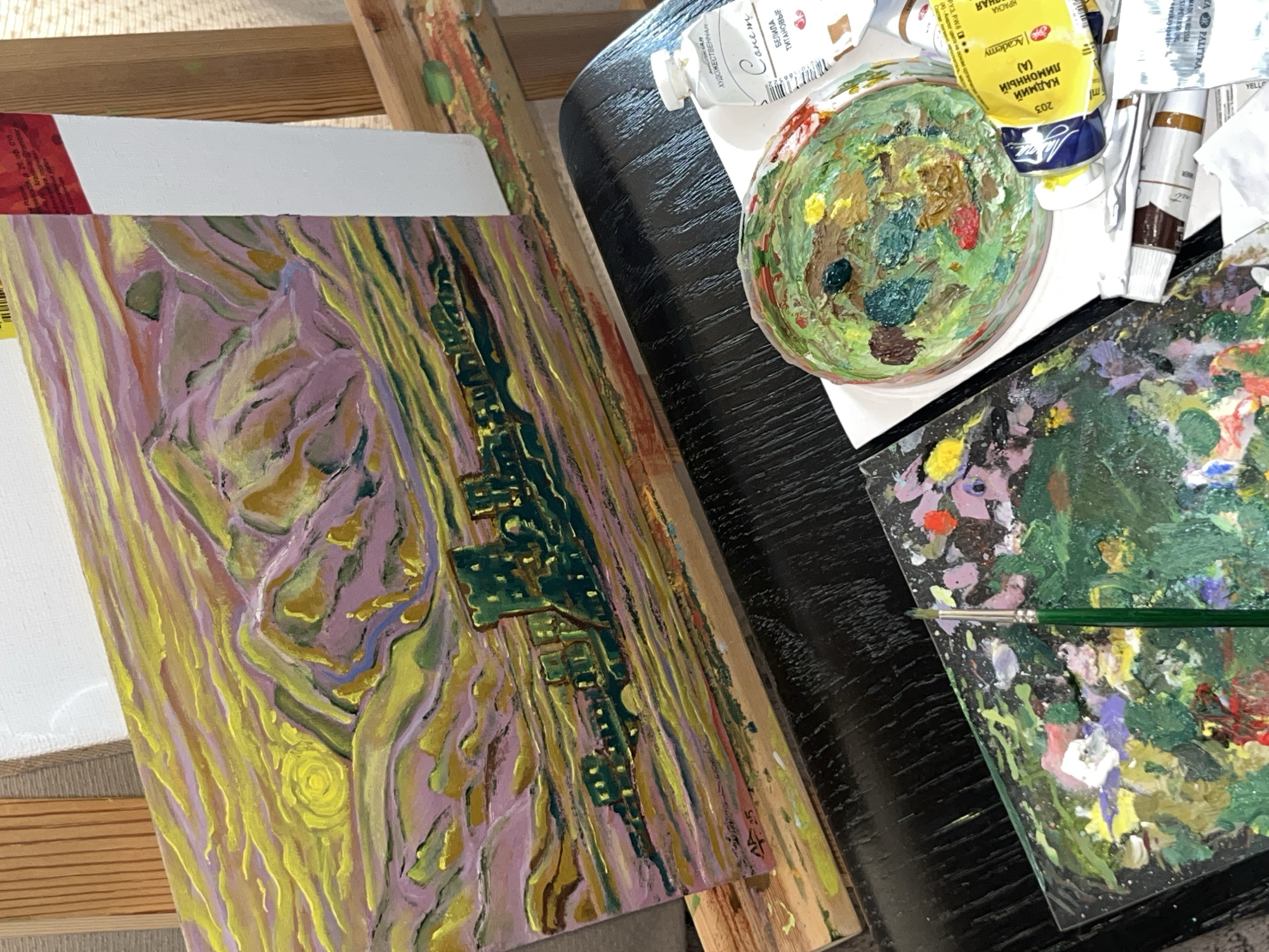 The image presents a tabletop still life in an art studio setting. It focuses on the artist's workspace and materials.
Subjects and Objects:
* A canvas with an unfinished landscape painting in progress, featuring purple, yellow, and green hues. It depicts what seems to be an abstract interpretation of a city view with a river.
* A color palette, likely containing a mix of oil paints, sits next to the canvas.
* Tubes of oil paints are present, including white and yellow paints with Cyrillic text. The yellow one mentions "КАДМИЙ ЛИМОННЫЙ (A)" and the white says "БЕЛИЛА ТИТАНОВЫЕ". These texts indicate Russian origin.
* Two palettes with mixed paints sit on the surface.
Environment:
* The setting is an art studio or workspace, evident from the presence of art materials.
* The tabletop is dark and potentially made of wood.
Actions and Interactions:
* The presence of the canvas, paint tubes, and palette suggests an artist is in the process of creating a landscape painting.
* The location is Krasnoyarsk, Russia, judging by the Cyrillic writing on the tubes.