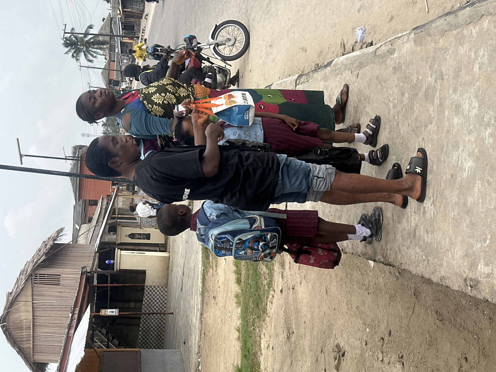 In Badagry, Nigeria, a group of people stand on a concrete sidewalk adjacent to a dirt road. A woman wearing a blue and green patterned dress is positioned in the center, holding a small shopping bag. To her left, a boy in a maroon uniform and a Captain America backpack stands, with a red bag at his feet. Another person, partially visible on the far left, wears a black t-shirt and denim shorts. Further to the right, a motorcycle with a rider is on the road. In the background, buildings with corrugated metal roofs and wooden structures are visible, along with a palm tree. The sky is overcast.
