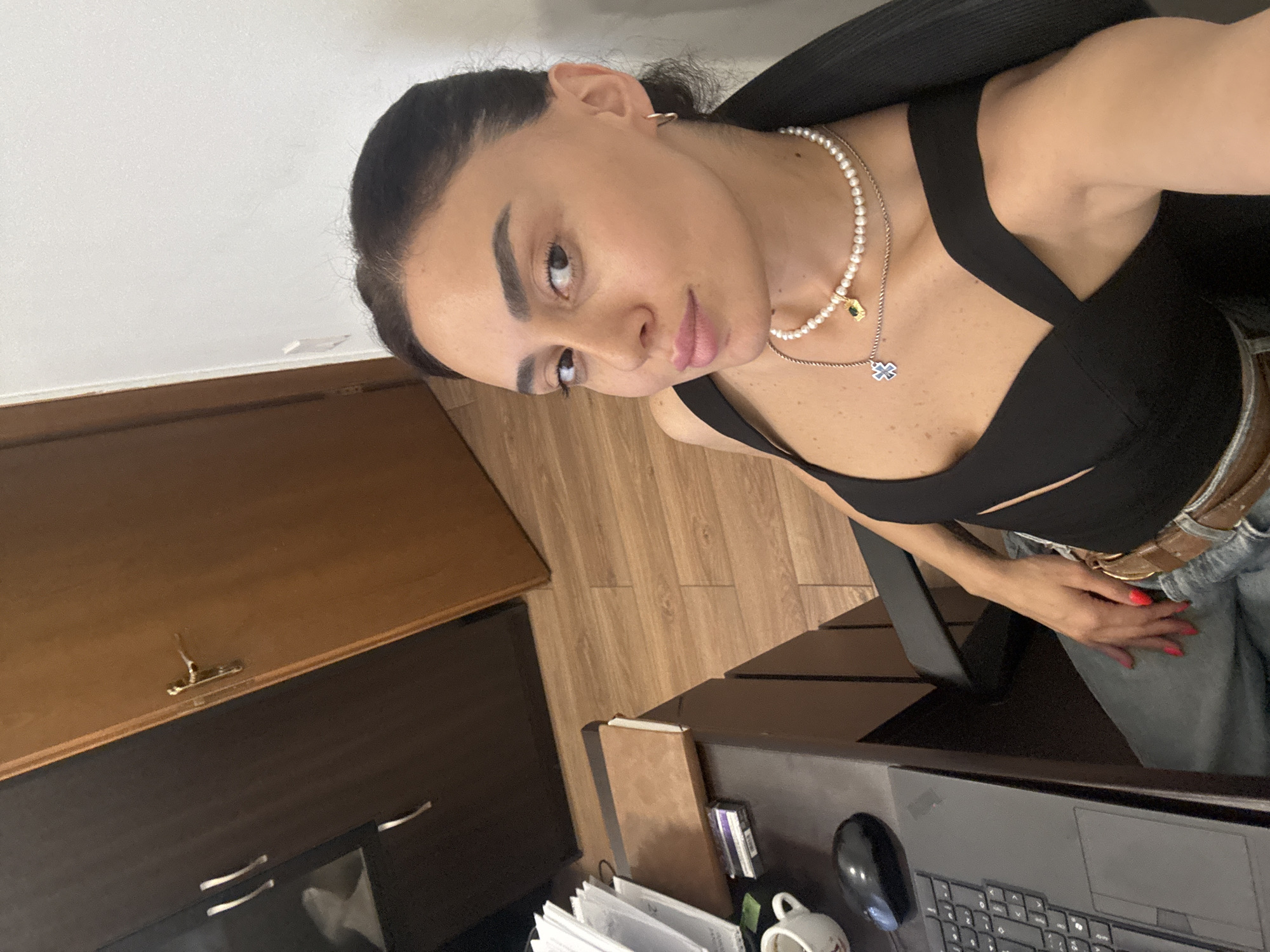 A medium shot photo shows a woman sitting at a desk in what appears to be an office or study. She is wearing a black top, jeans, and jewelry including a pearl necklace with a pendant. She has dark hair pulled back and is looking towards the camera. On the desk are a laptop, a mouse, a coffee mug, and some papers. In the background, there is a dark cabinet, a wooden door, and a white wall. The floor is wooden. The scene is indoors and appears to be in an urban environment. There are no obvious indicators to specify the exact city, however, the presence of a dark wooden cabinet with chrome handles and a light wooden door with a brass handle are common features in homes and offices. The overall lighting suggests it is daytime.